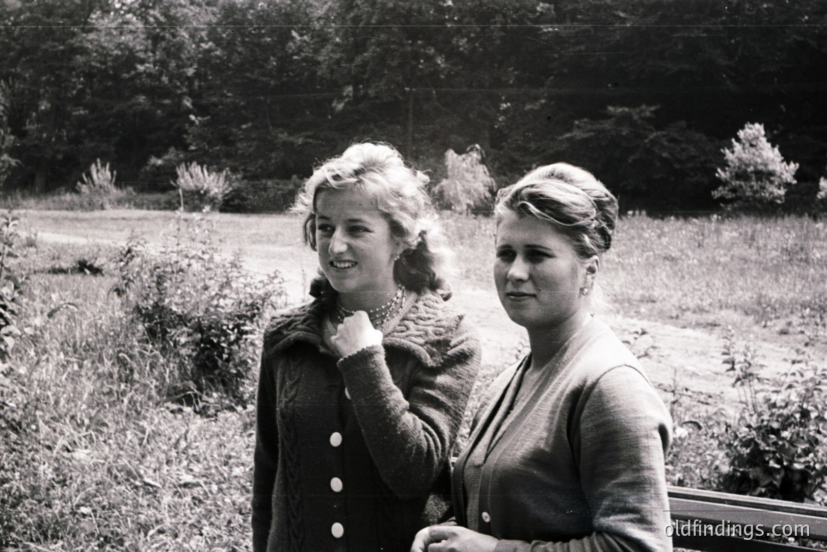 Two young women in cardigans stand near a park bench, gazing off-camera. Their hairstyles and clothing suggest a mid-20th century style, likely the 1950s or early 1960s. The background shows dense foliage and a grassy area. A candid, natural moment captured in black and white.