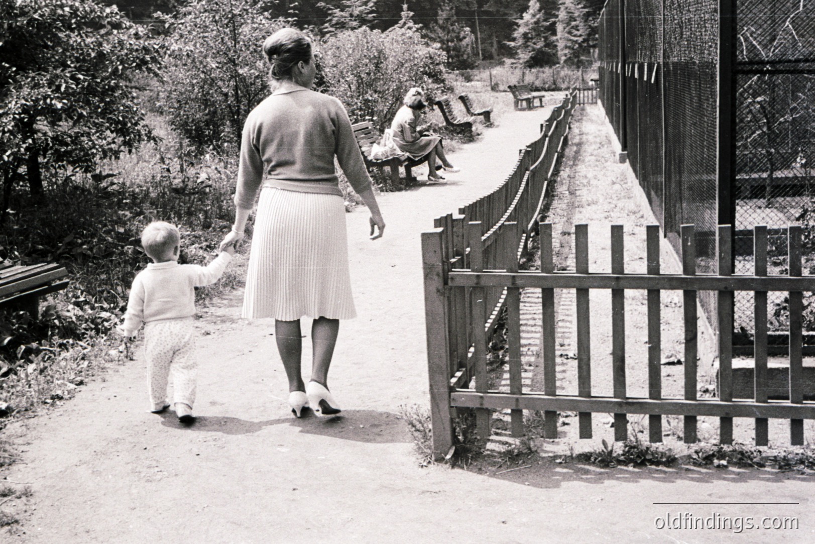A woman and toddler walk along a paved path, hand-in-hand, next to a wooden picket fence. A seated figure rests on a bench visible in the distance. Woman wears a pleated skirt and heels. Appears to be a park or zoo setting. Likely 1950s-1970s. Stock photo potential: family, childhood.