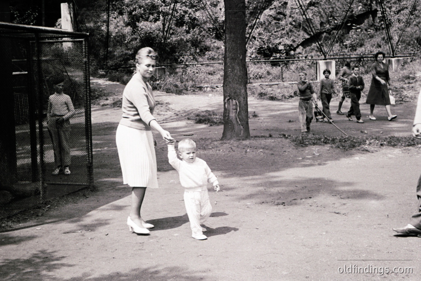 A mother and toddler walk hand-in-hand on a gravel path, likely in a park or zoo. The child, in overalls, takes unsteady steps. A fenced enclosure and other park visitors are in the background. Appears to be mid-1960s, vintage snapshot style.