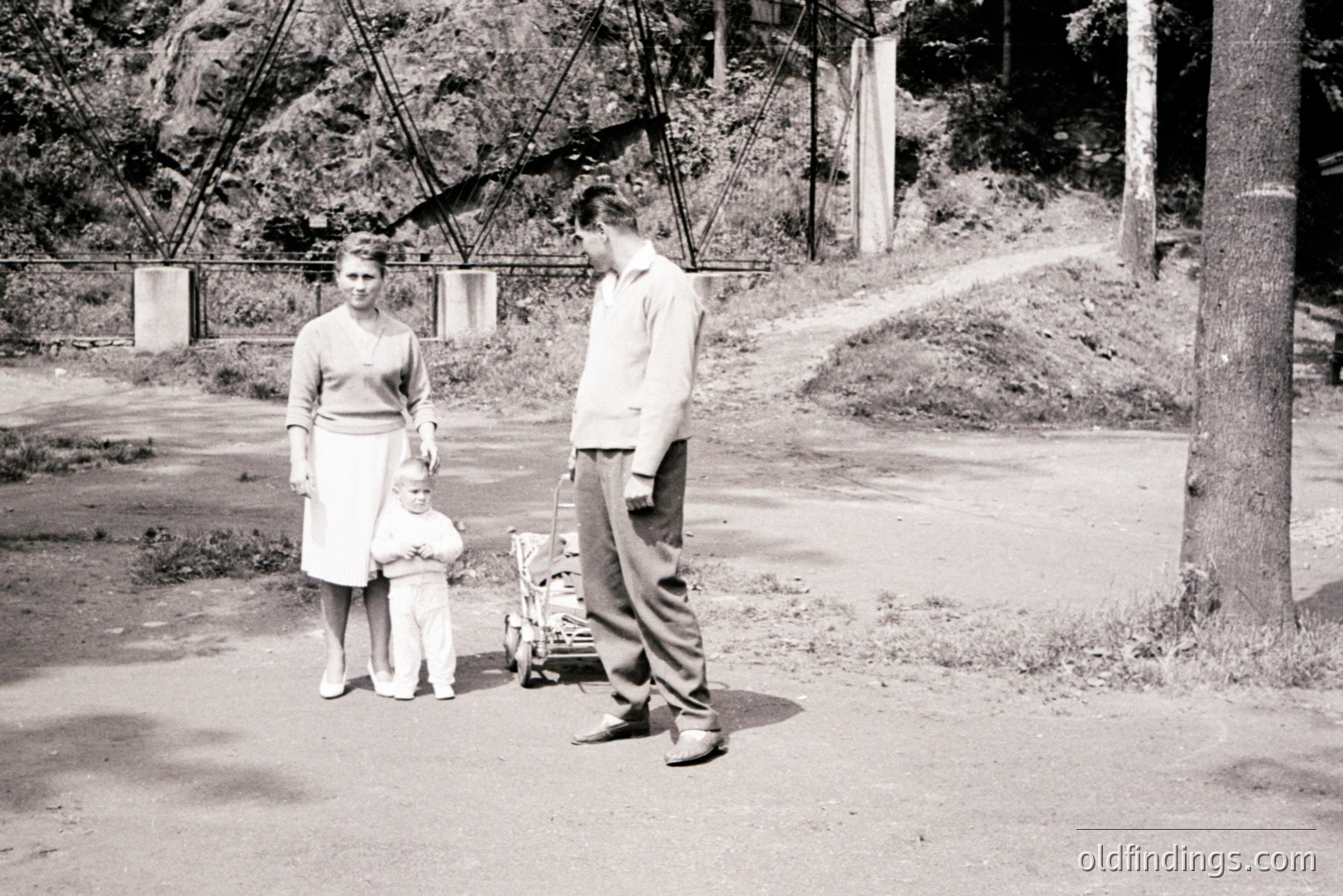 A young couple stands with a toddler outdoors, near a substantial bridge or viaduct structure. The woman wears a modest dress; the man, workwear. A small, wheeled plaything sits between them. Likely 1950s, potentially a vacation or outing. Subject matter valuable for family history or design reference.