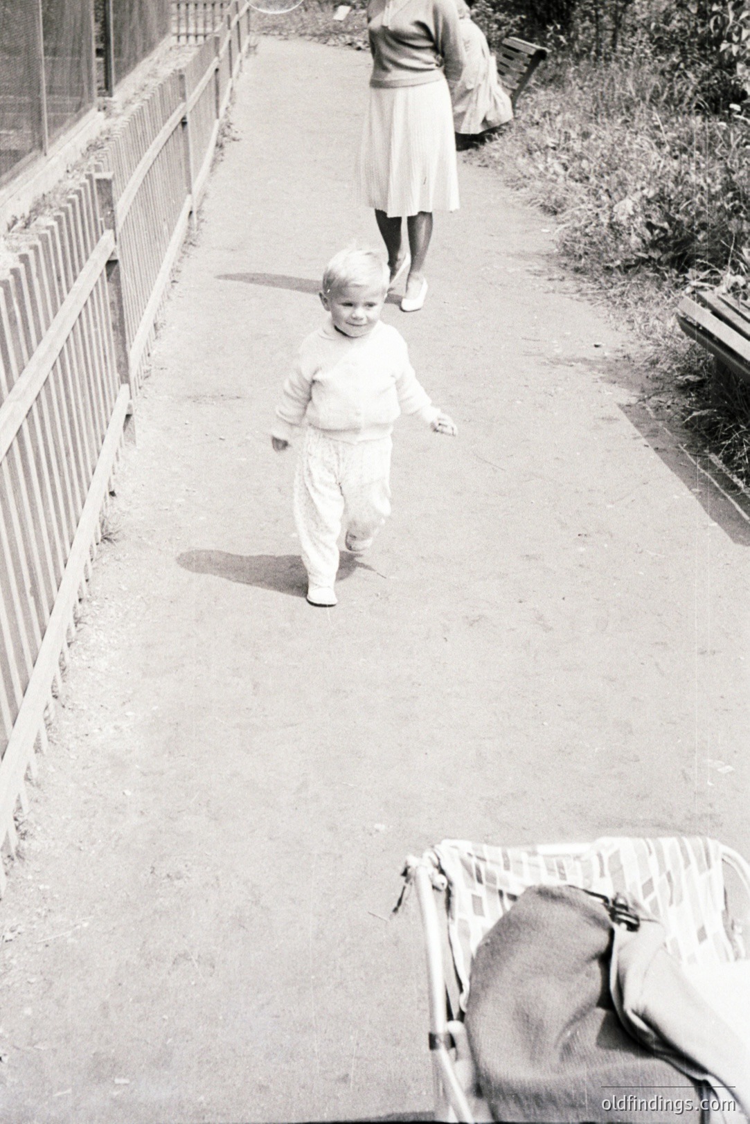 A young boy in a sweater and pants walks along a paved path. An adult in a skirt and carrying a bag walks away in the background. A stroller sits abandoned near the curb. Likely mid-20th century, evoking a nostalgic, everyday family scene.