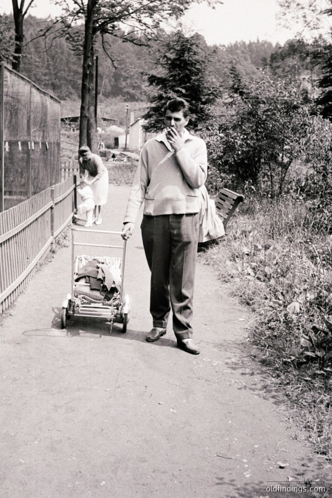 A man in a sweater and trousers strolls along a rural road, pushing a vintage baby carriage. A young girl in a white dress is visible in the background. Likely 1960s-70s, this scene evokes a quiet, domestic moment. The composition suggests an everyday, ordinary life.