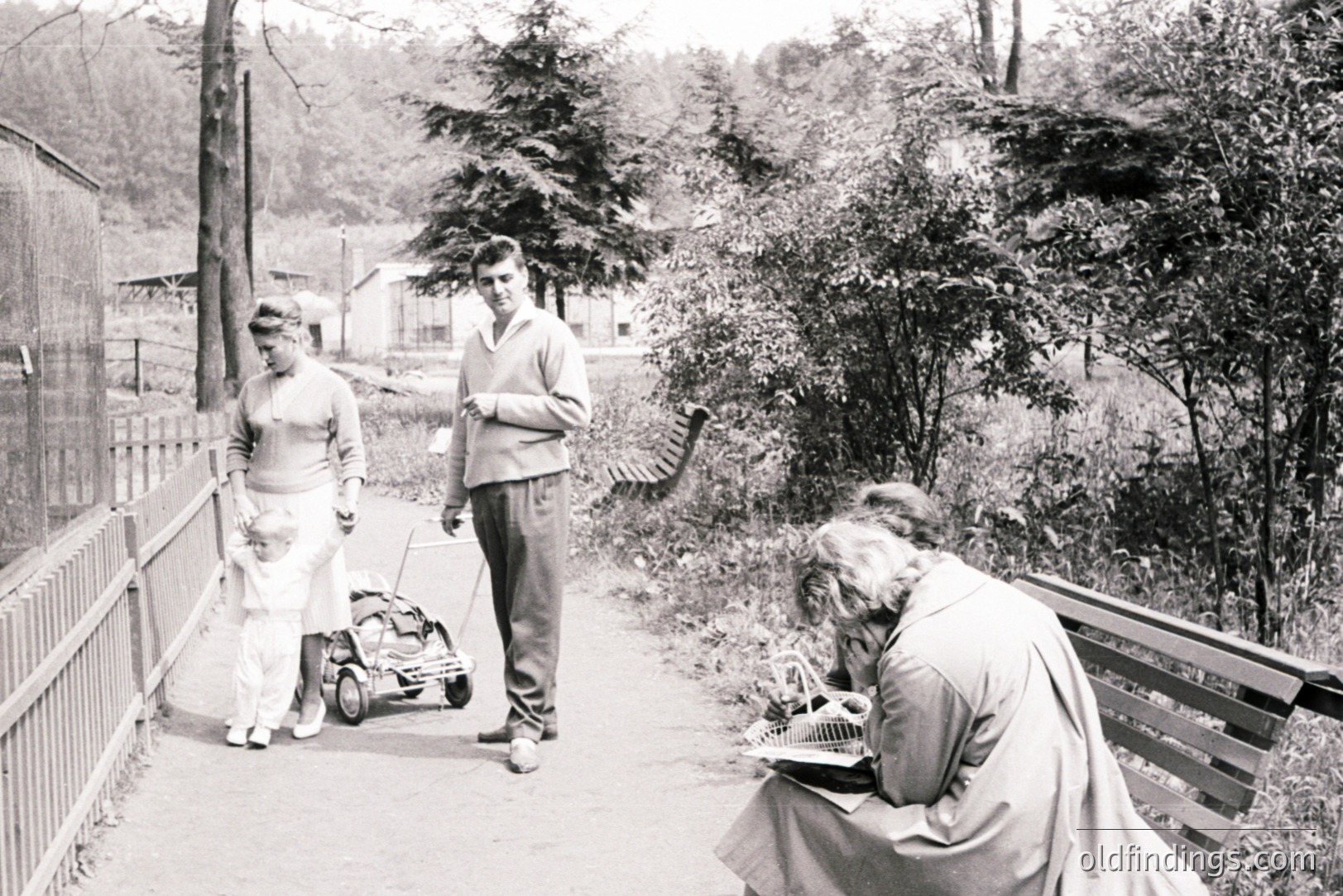 A young family strolls along a pathway, with a baby in a vintage pram. A woman sits on a bench, sketching in a notebook. Architectural details suggest a mid-century European park setting. Likely 1950s-1960s. Strong potential for commercial use depicting family life.