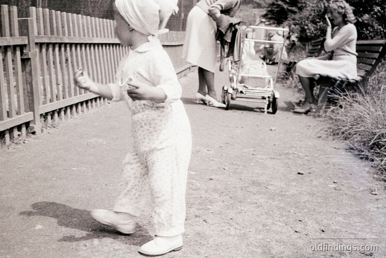 A young child, clad in white overalls and a matching headscarf, walks away from the viewer, casting a prominent shadow. Two women are present - one pushing a stroller, the other seated on a bench, both in 1930s-style attire. Appears to be a domestic scene, possibly a garden or estate.