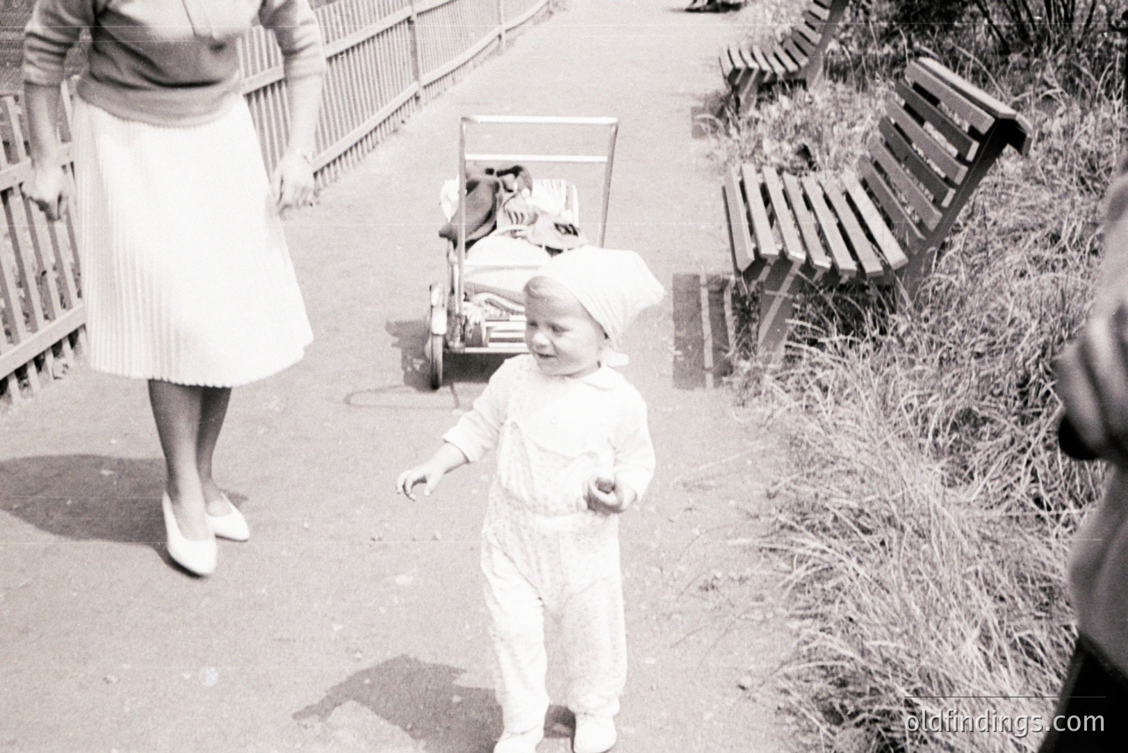 A young child in overalls and bonnet walks forward, seemingly delighted. A woman in a pleated skirt and heels walks behind, pushing a vintage pram. Park bench & foliage are visible. Likely a family outing. Evokes a sense of mid-century domesticity.