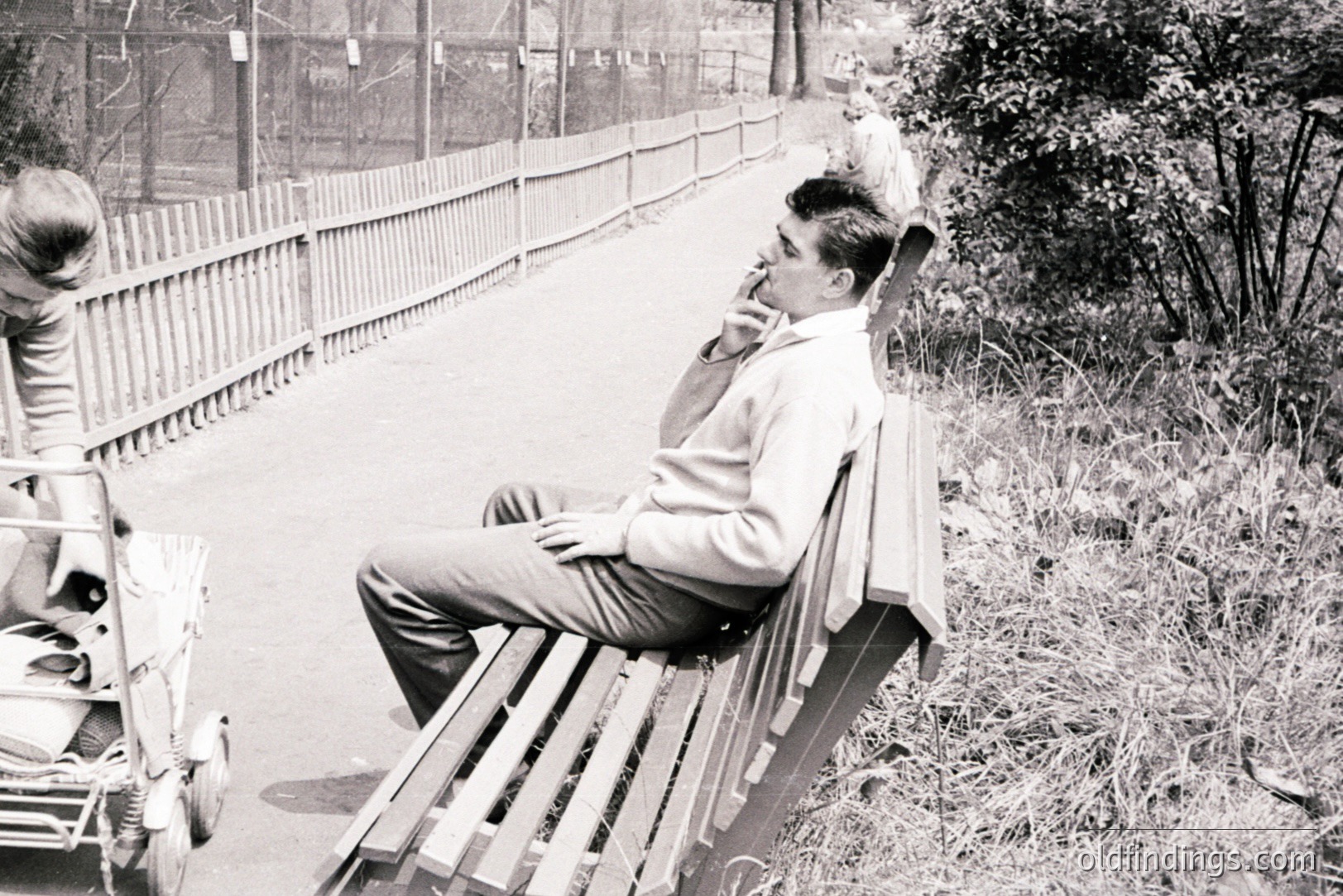 A young man in a sweater and slacks sits pensively on a wooden bench. A partial view of a woman and a vintage scooter are visible to the left. A low fence and foliage line a paved path. Likely 1960s, candid street scene. Suitable for design, vintage advertising or historical stock.