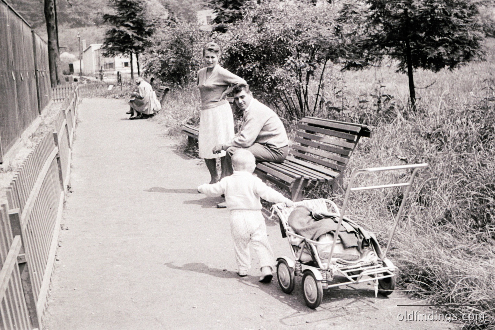 A young boy pulls a vintage baby stroller along a paved path. A couple stands nearby, observing him. A seated person is visible further down the path. Likely a 1960s family scene, suggesting nostalgic appeal.