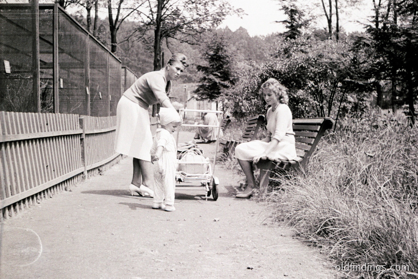 A young child, likely a toddler, stands near a vintage pram on a paved path. Two women, possibly a mother and nanny, are present, one attending the child, the other seated on a bench. Likely a public garden or park setting with mature trees in background. Appears to be a candid moment, circa 1930s-1950s.