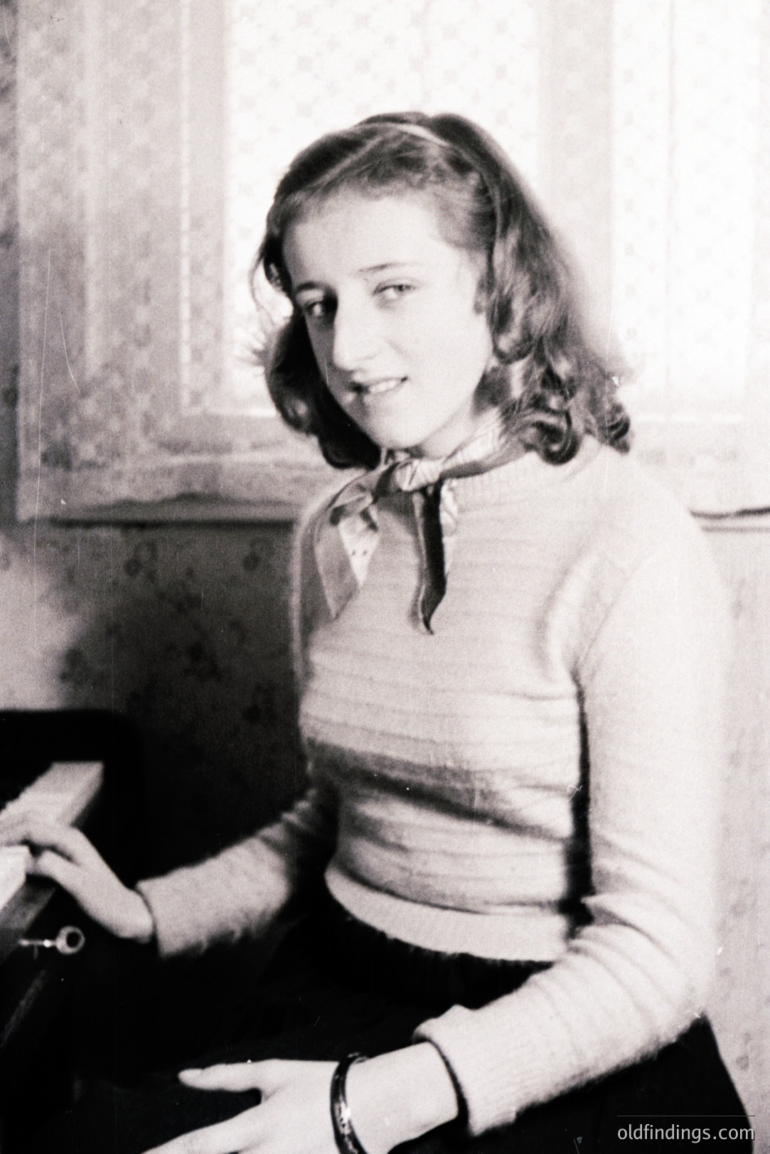 Young woman in a ribbed sweater and dark skirt, seated with a slight smile. A patterned scarf adorns her neck. The background includes a window and patterned wallpaper, typical of mid-century interiors. Likely a family portrait, c. 1950s-1960s. A valuable reference for vintage fashion & interiors.