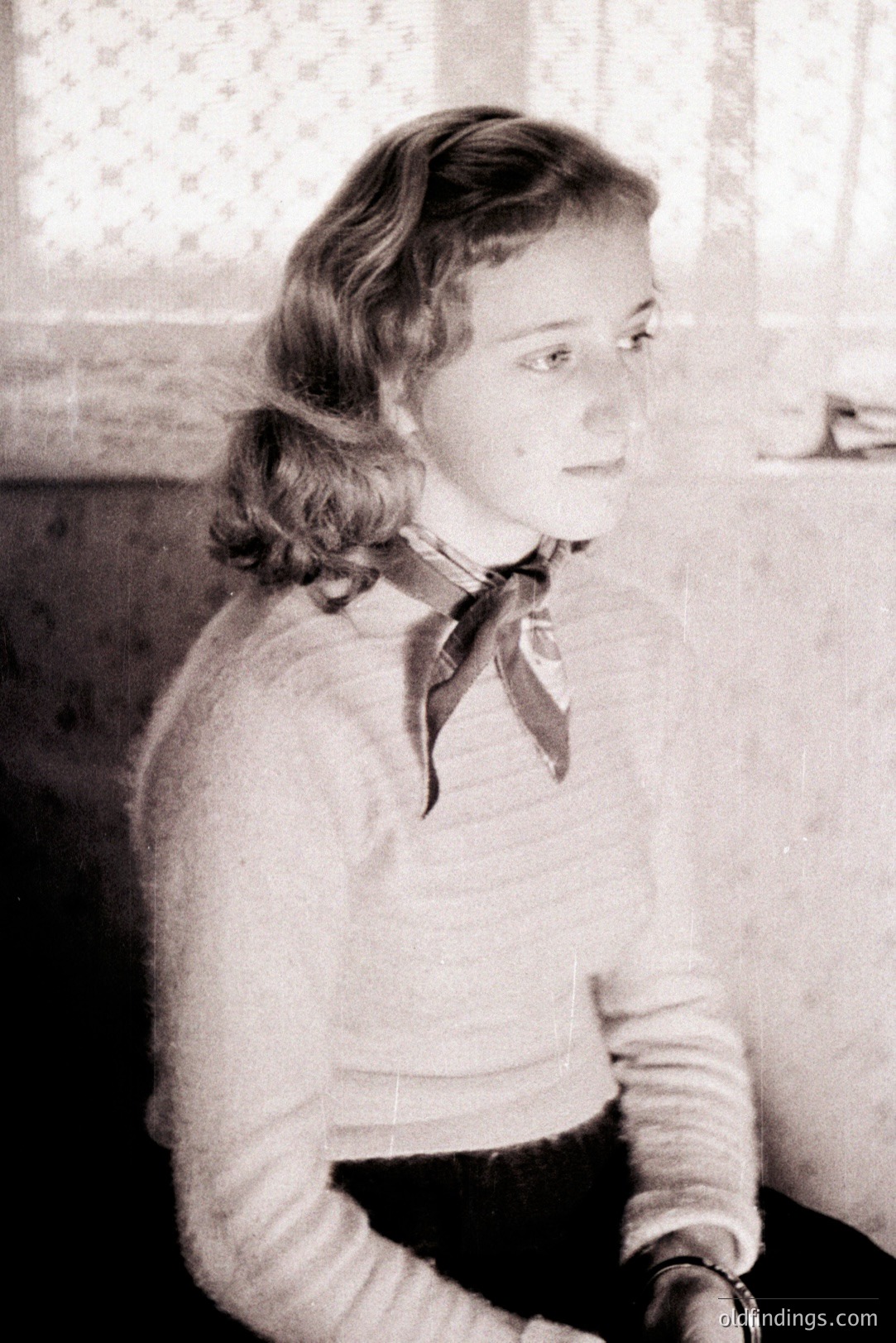 A young woman with styled hair and a dark scarf sits indoors, captured in a medium shot. She's wearing a textured sweater and dark skirt, gazing slightly off-camera. The image, with its grainy quality, suggests a portrait from the mid-20th century, likely the 1940s or 50s. Soft lighting highlights a simple domestic scene.