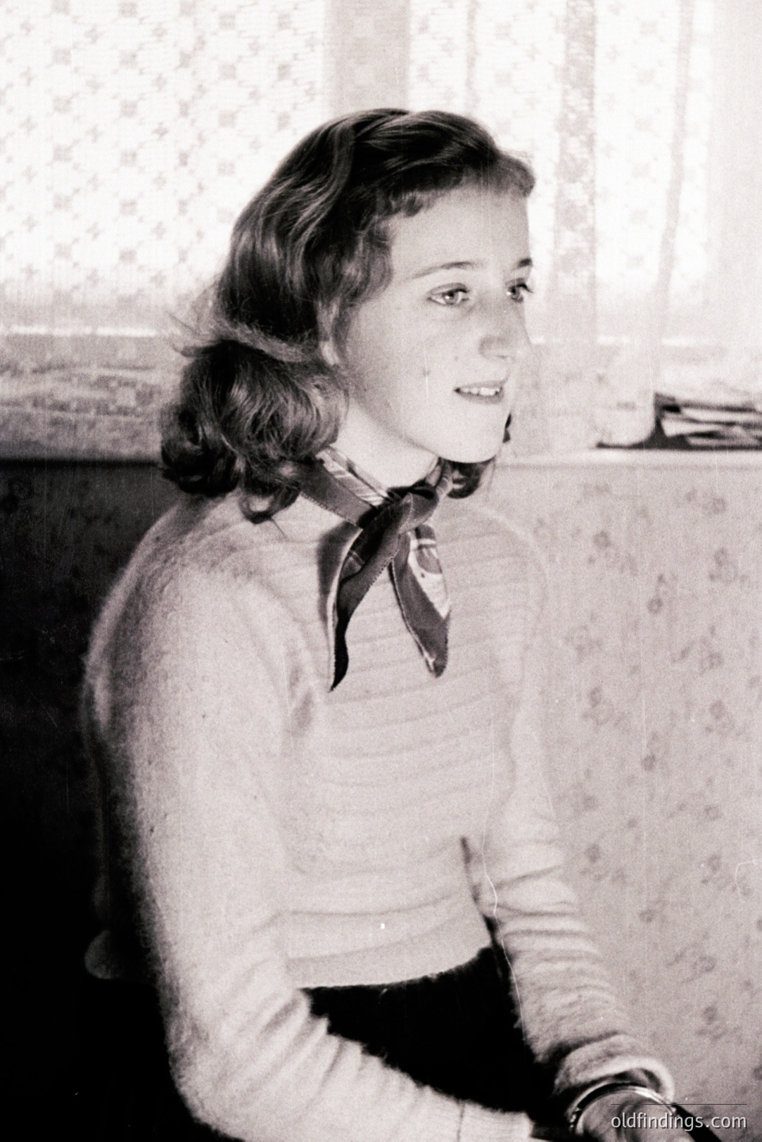 Young woman in a textured sweater and dark skirt, a silk scarf knotted at her neck. Seated indoors, looking over her shoulder with a slight smile. Likely a portrait from the 1950s, reflecting post-war fashion and domestic settings. Wallpaper visible behind. A classic, timeless aesthetic.