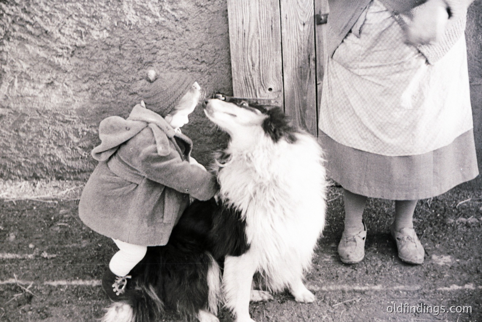 A young child in a hooded coat reaches up to pet a fluffy black and white dog, posed near a weathered wooden door. The woman beside the child wears a simple, homespun dress and work shoes, suggesting a rural setting. Likely a snapshot from the 1950s-1970s, capturing a tender moment. Could be valuable for illustrating family life or vintage aesthetics.