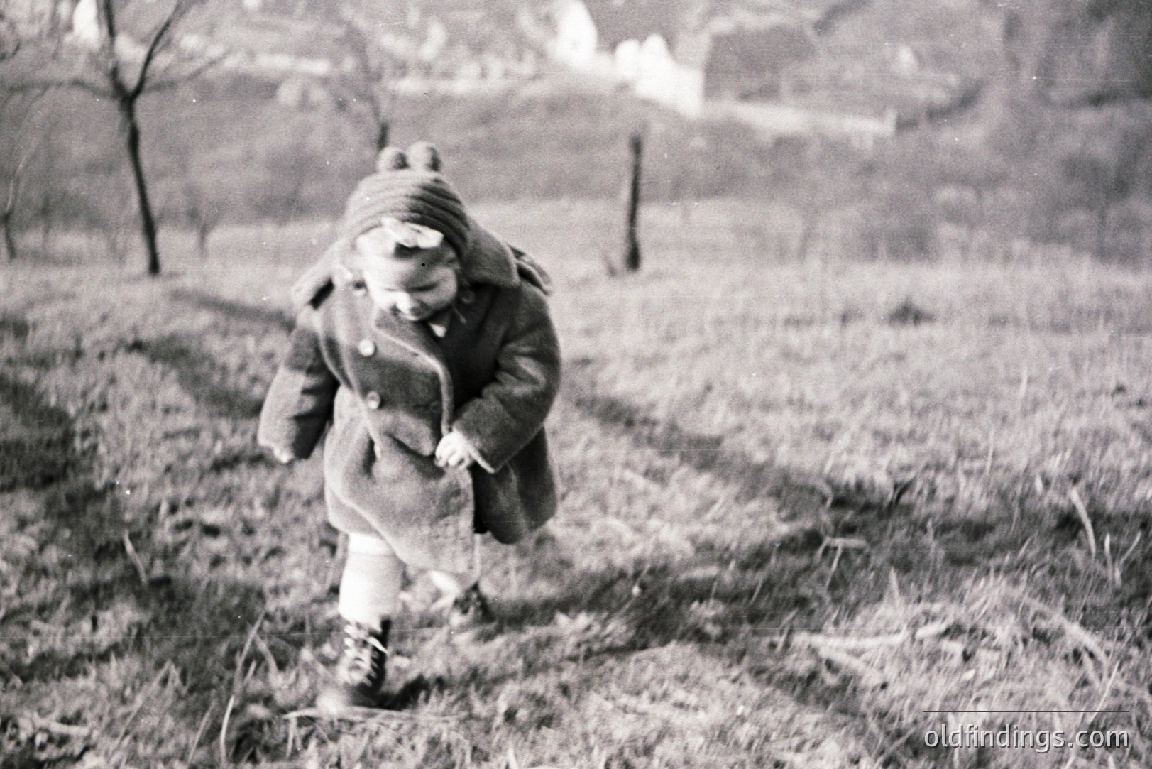 A young child, bundled in a wool coat and hooded hat, runs down a grassy path. The scene appears to be a rural landscape, with trees and a building visible in the background. Likely a candid snapshot, evoking a nostalgic, mid-century feel.