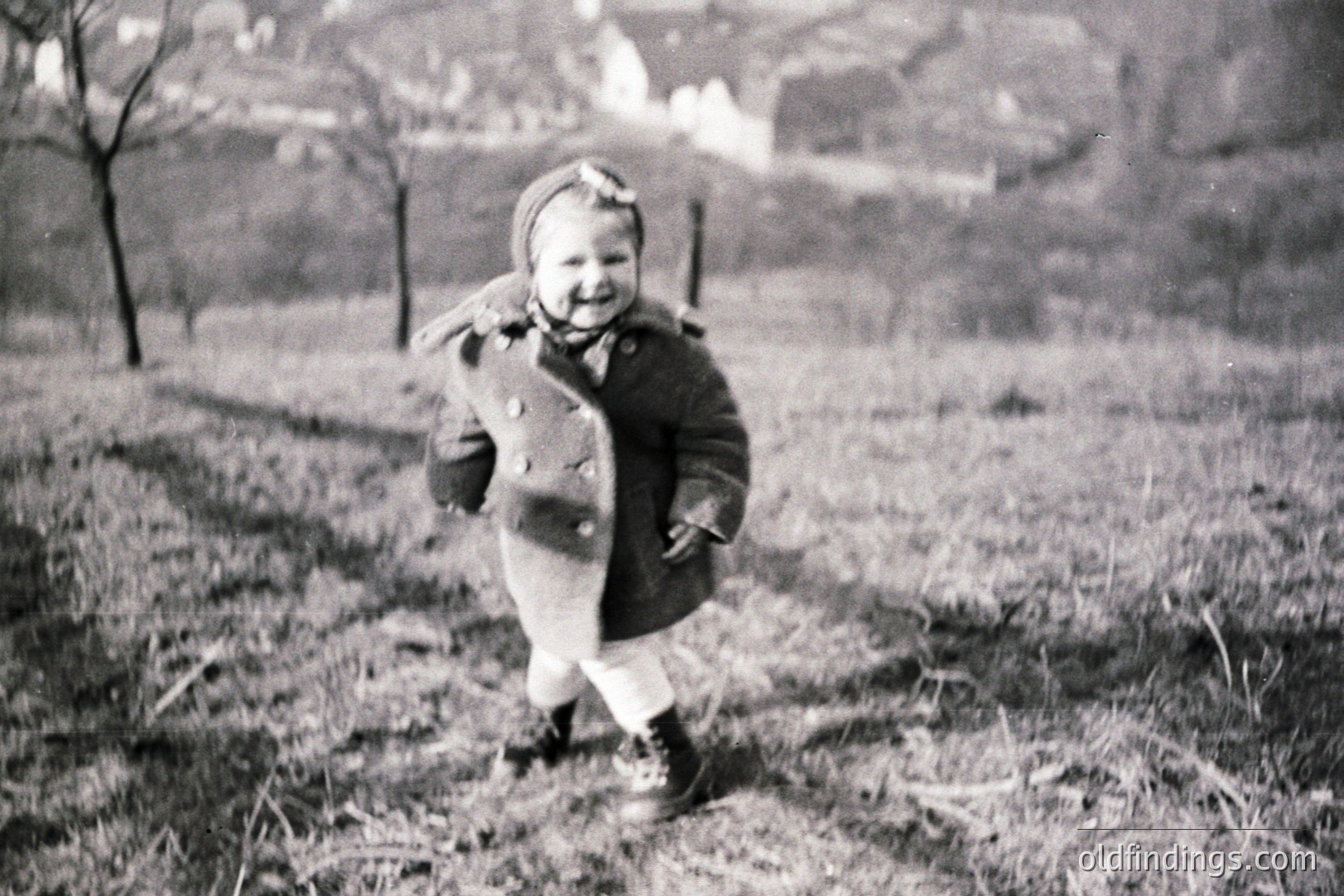 A young child, likely a toddler, runs toward the camera wearing a heavy wool coat, a floral headband, and laced boots. Set against a backdrop of bare trees and a distant building. Appears to be a rural landscape, possibly taken in the 1940s or 50s. Charming, candid family portrait.