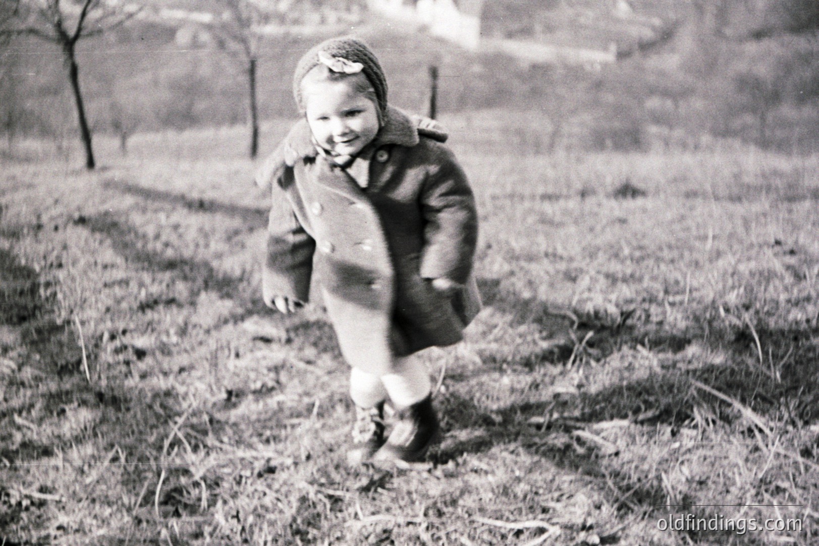A young child in a wool coat and bonnet runs across a grassy field, captured in a moment of playful motion. The vintage photograph, likely from the 1950s, shows a simple, rural scene, possibly a family outing. The style suggests stock photo or archival family documentation potential.
