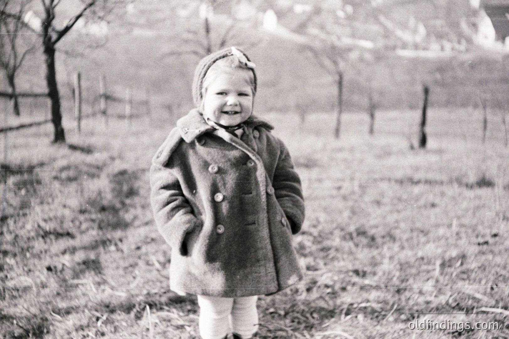A young child, likely a toddler, stands in a grassy field wearing a heavy wool coat and a decorated hat. The setting appears rural with a blurred background of trees and a fence. Likely a candid portrait, capturing a moment of childhood joy. Likely 1950s-1960s.