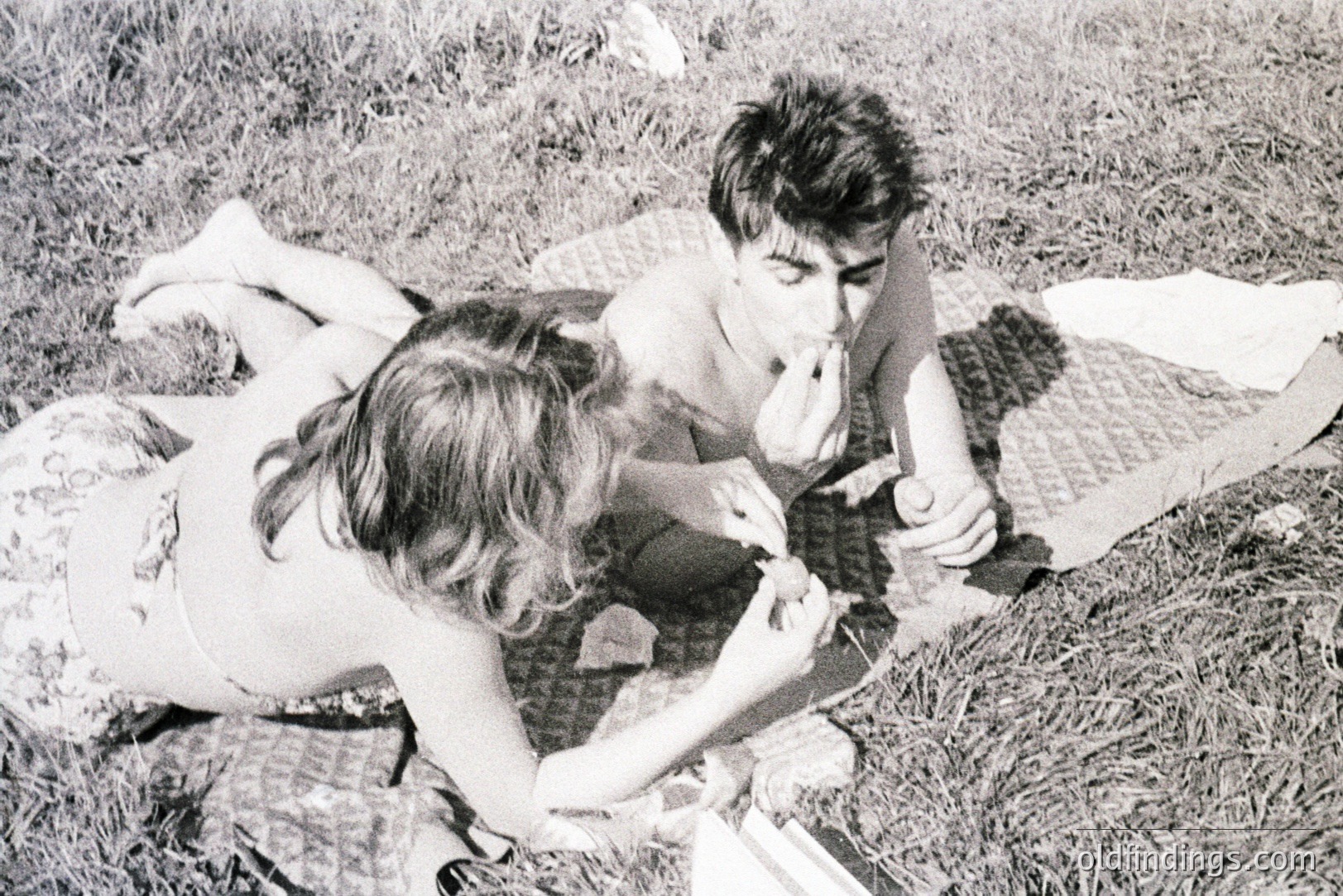 Young couple enjoys a picnic on a grassy lawn. The woman, in a floral print top, offers food to the shirtless man. A patterned blanket and scattered tableware suggest a casual, relaxed setting. Likely 1960s style. Appealing for retro lifestyle imagery.