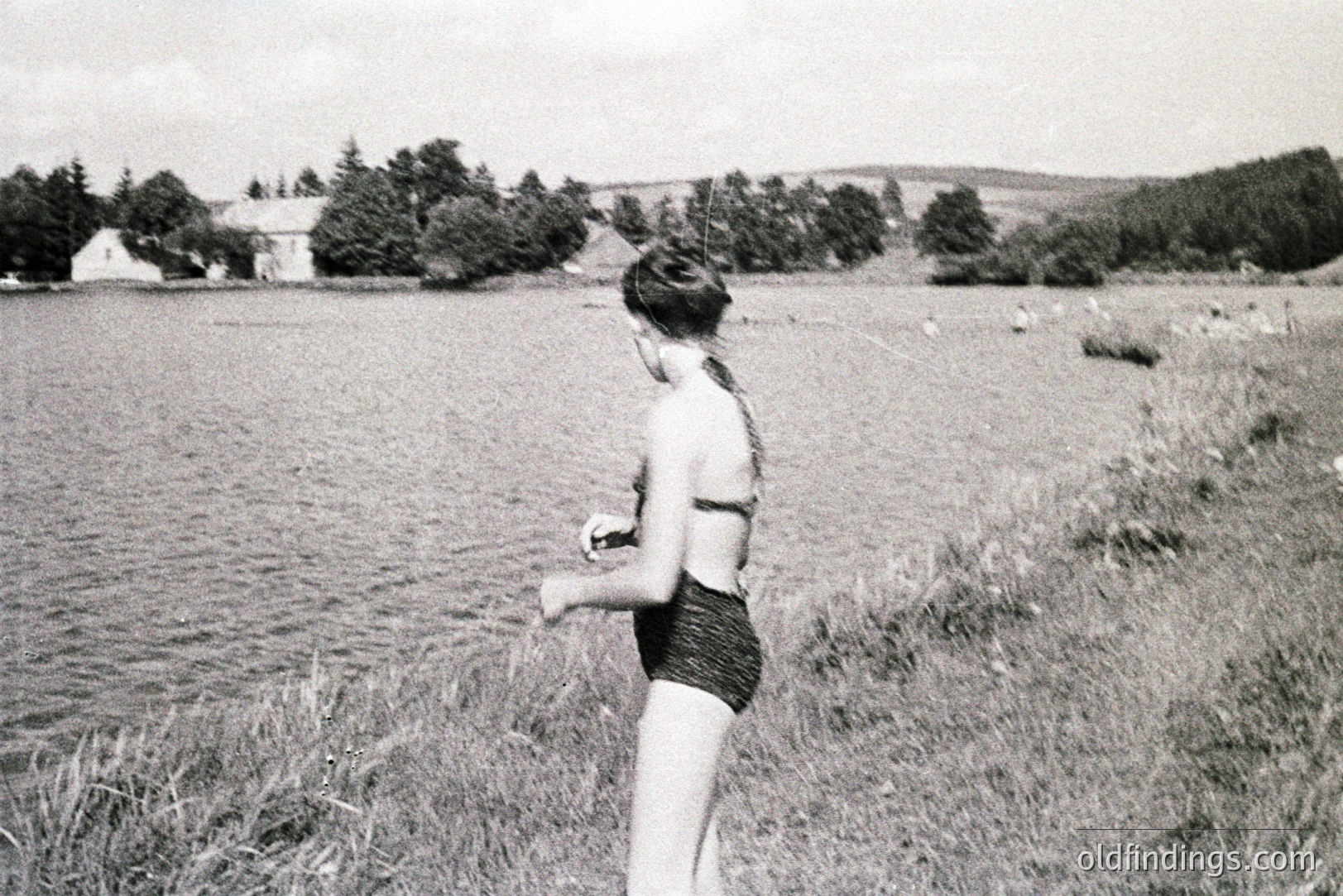 A young woman, viewed from behind, stands near a body of water, likely a lake or reservoir. She wears a textured, dark-colored bikini. Lush greenery and trees form a backdrop; a building is partially visible. Grainy texture suggests a vintage film photograph, possibly 1960s or 1970s. A serene, natural setting.