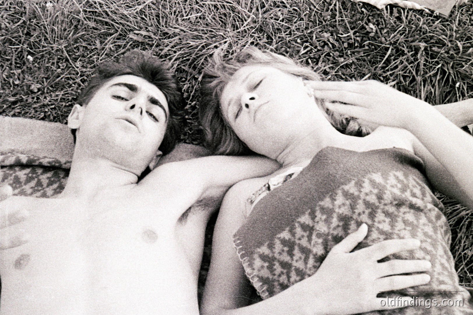 A black and white photo depicts two young people lying closely in grass, seemingly asleep. The male figure is shirtless, and the female wears a patterned top. Composition utilizes a high-angle perspective. Likely a personal snapshot or amateur photography.