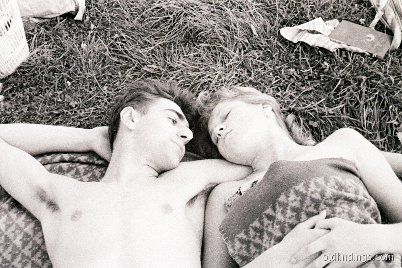 A young couple lies intertwined on a grassy area, heads close together, eyes closed; the man shirtless, the woman on a patterned blanket. Suggestive of relaxation and intimacy, captured in a candid, grainy style. Likely a 1970s snapshot. Potential for stock photography or design reference.