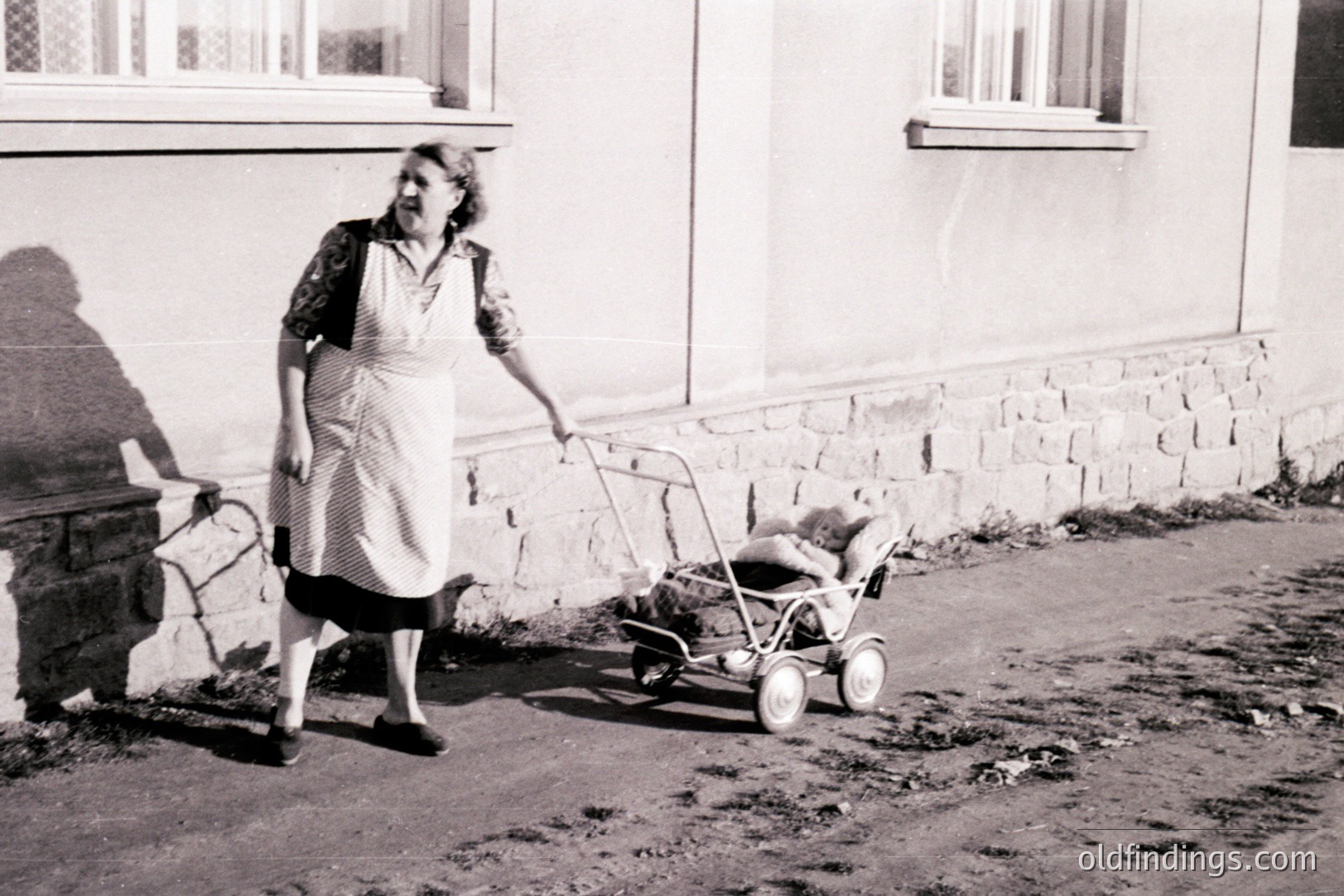 A woman in a dark dress and apron pushes a vintage, wire-mesh stroller along a paved path alongside a stone building. The photo appears to be from the 1940s-1950s, likely depicting domestic life. A long shadow stretches across the pavement. Evokes a sense of everyday routine and simple living.