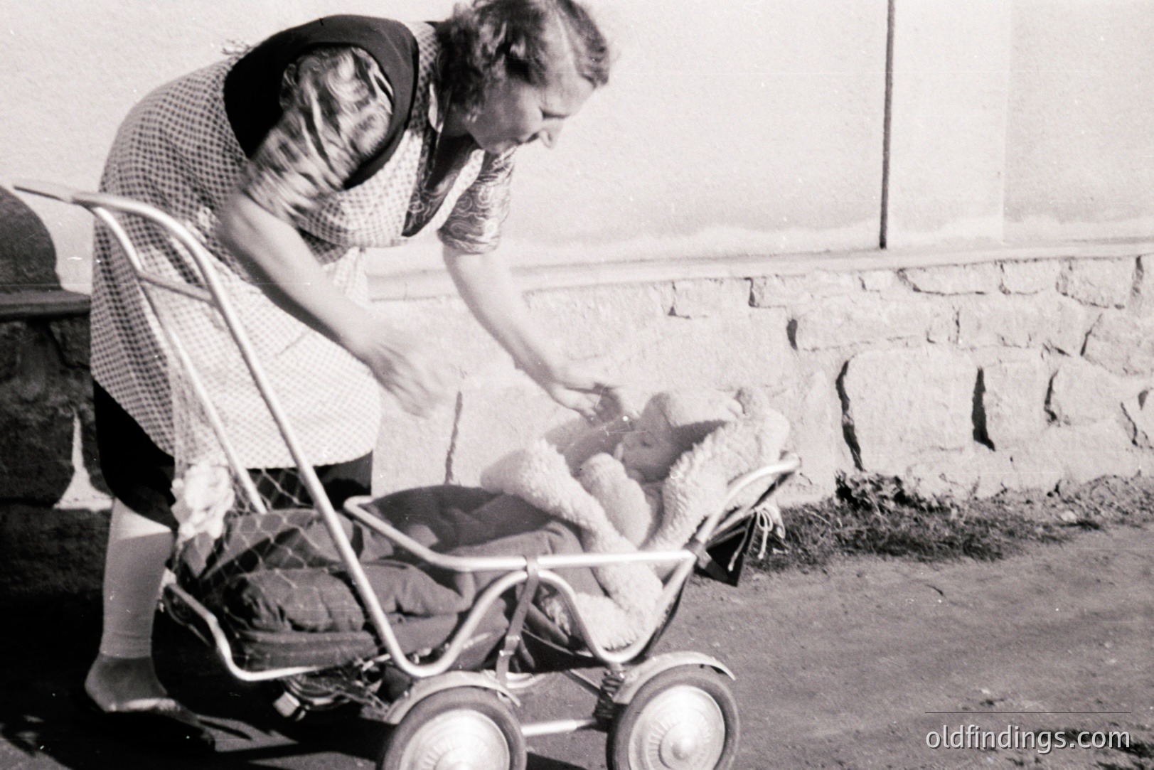 A young woman in a patterned dress and apron tends to a baby in a vintage, wheeled stroller featuring unusual spoke-style wheels. A large teddy bear sits alongside the infant. Likely taken outdoors, possibly near a coastal location, judging by the stonework and flat horizon. Estimated 1950s-1960s. Nostalgic family moment.