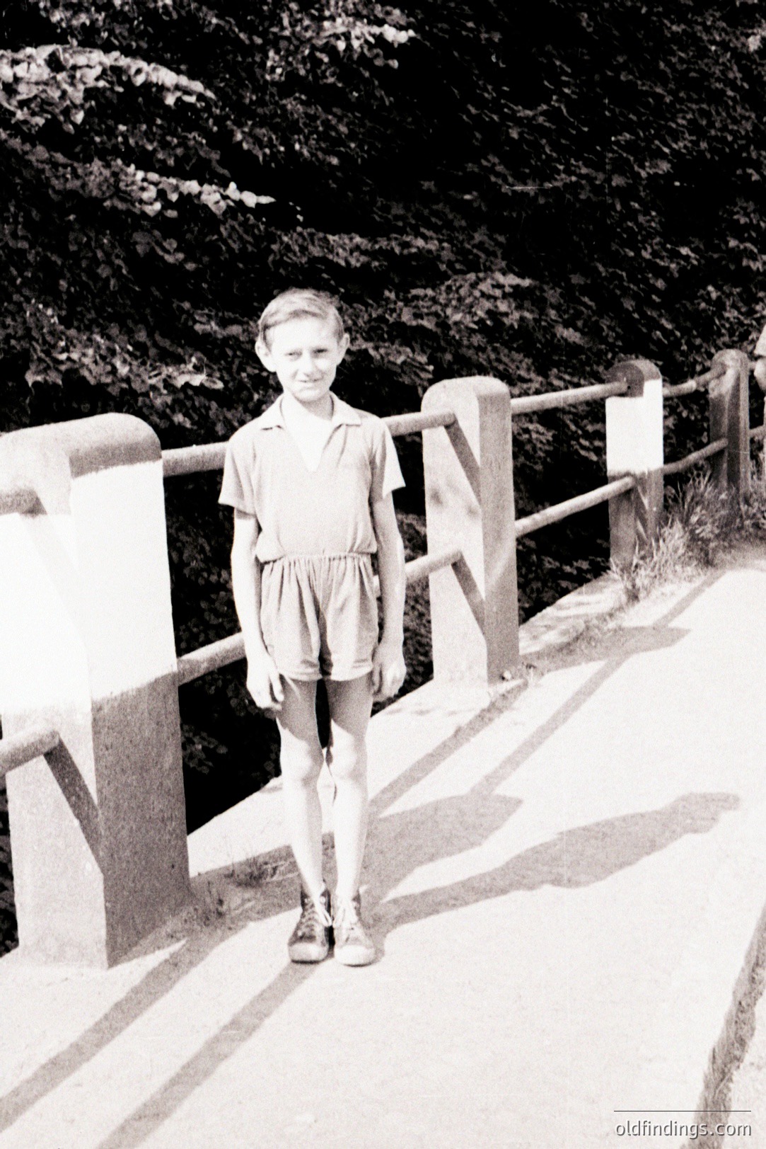 Young boy in a romper suit stands on a bridge with ornate railings, appearing mid-stride. The scene’s dark background suggests a wooded location. Clothing and hairstyle suggest a mid-20th century, likely 1950s or 60s, setting. A candid portrait likely for family documentation.
