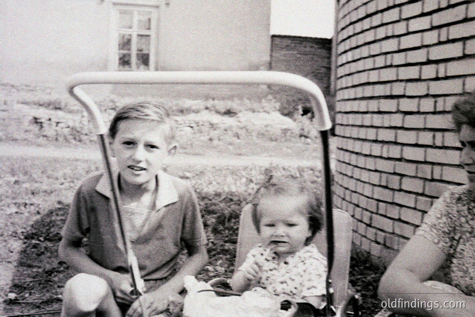 Black and white photo depicts a young boy seated beside a baby in a high-backed stroller. Brick wall and simple architecture suggest a post-war suburban setting, likely 1950s-1960s. Child's clothing and stroller design indicative of the era. Family snapshot with potential value for genealogy or historical research.