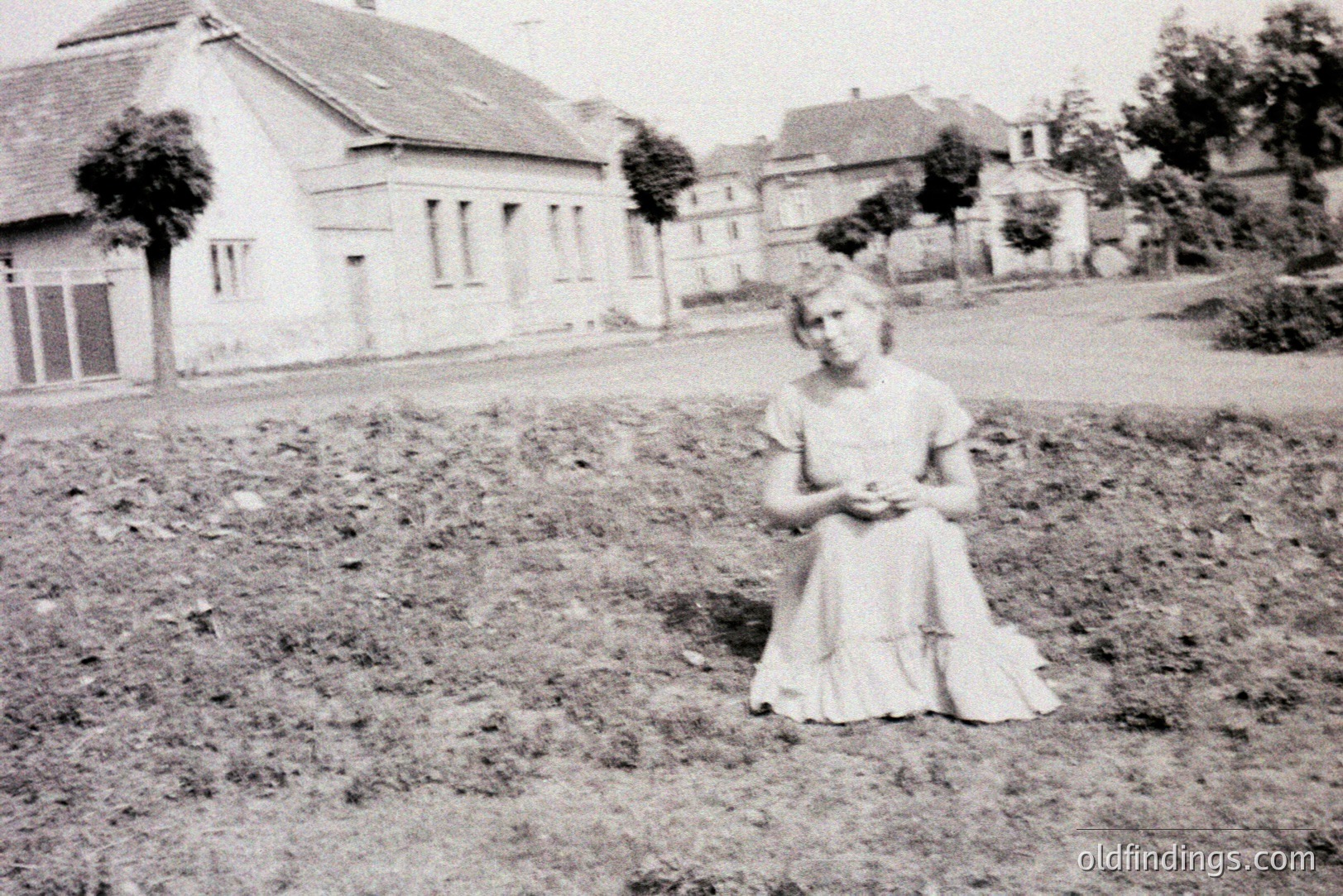A young woman kneels in a garden, wearing a modest, tiered dress. Background shows residential architecture with multiple houses and greenery. Appears to be a street scene, likely a suburban or rural setting. Possibly taken in the 1930s-1950s based on dress style.