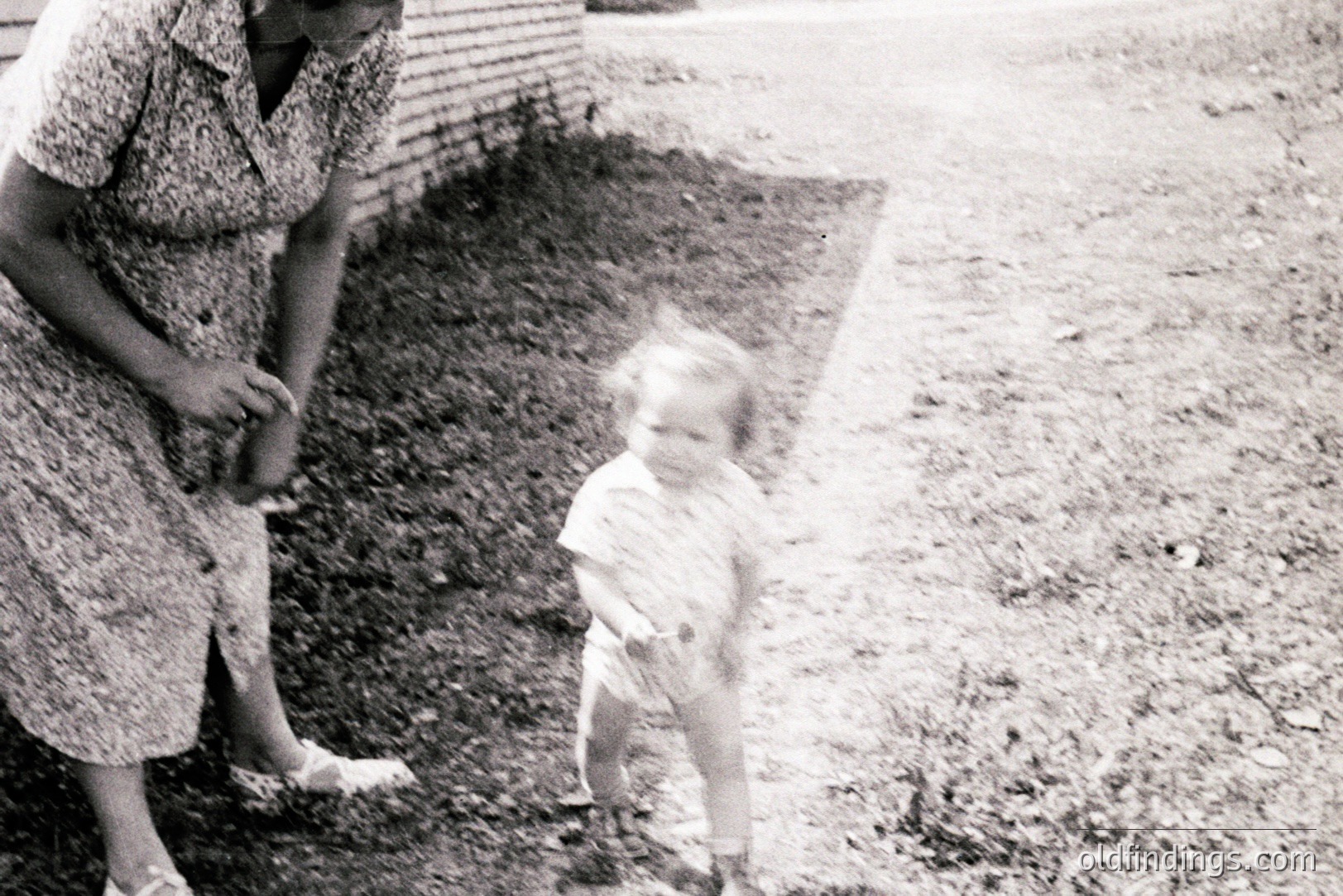 A young child in a diaper and short-sleeved shirt stands on a gravel path, appearing to run towards an adult woman in a patterned dress and sandals. Likely a candid family moment captured in the mid-20th century. The backdrop features a building with corrugated metal siding. A charming, everyday scene.