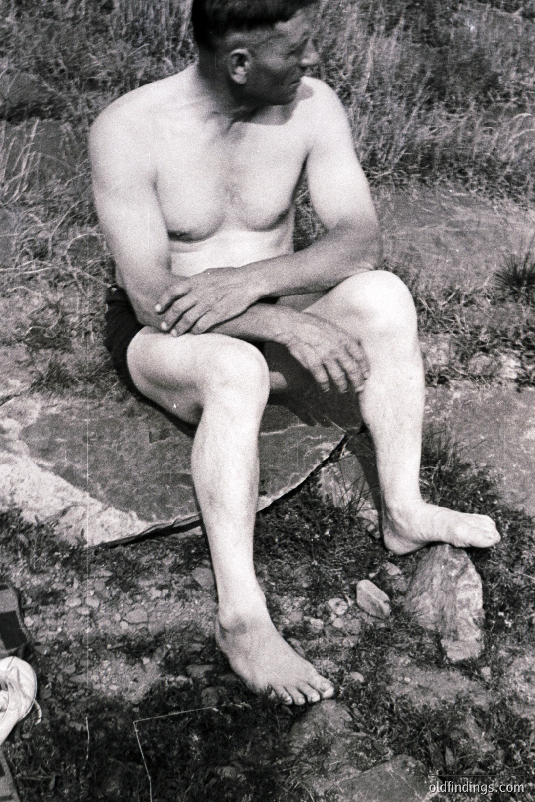 A seated, barefoot man, unclothed from the waist up, rests on a large, textured rock amongst sparse vegetation. The image is a grainy, monochrome, likely a vintage portrait study capturing detail in skin texture and musculature. Photographic style hints at a mid-20th century aesthetic. Location and ethnicity are indeterminable.