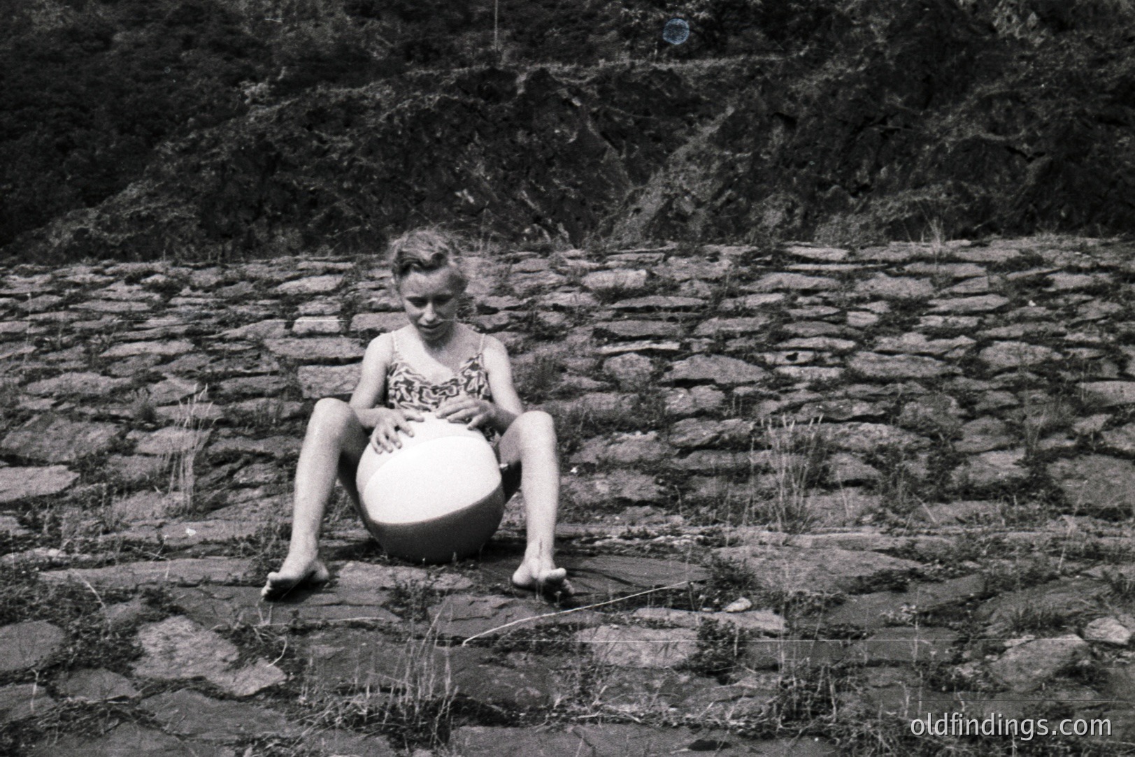 A young girl sits outdoors, cradling a round object (likely a ball) between her legs. She’s barefoot, seated on a grassy area adjacent to a stone wall. The black and white photography suggests a mid-20th century origin. Architectural detail and simple composition suggest documentary or candid style.