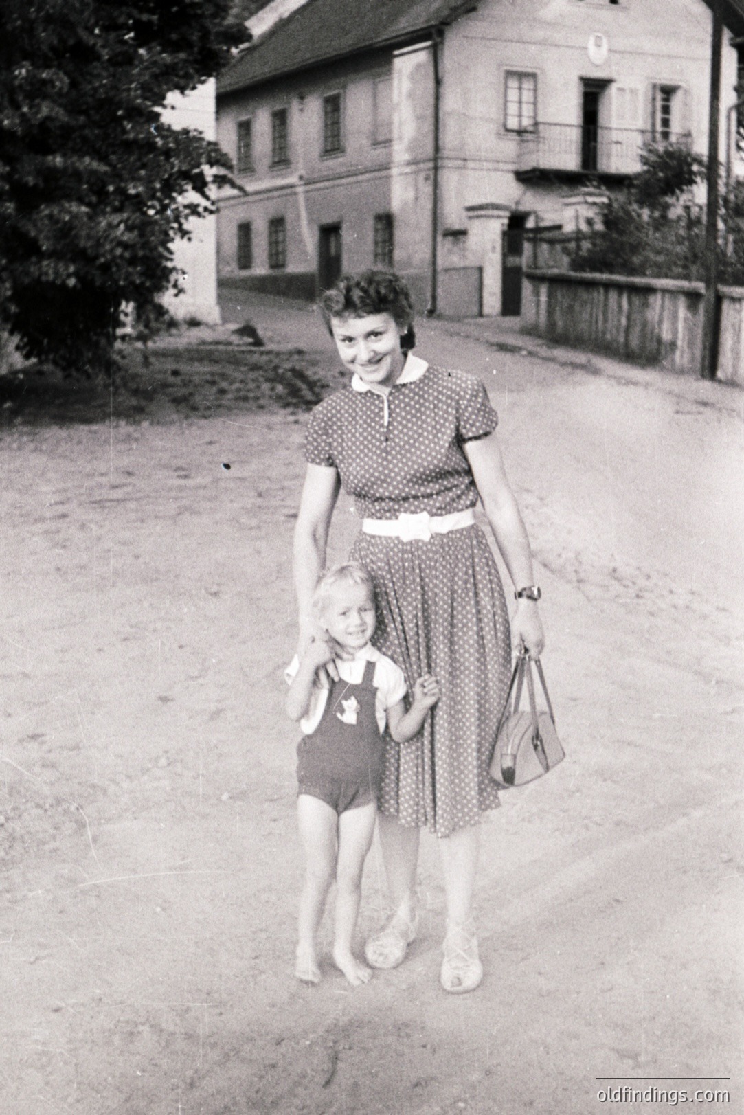 A mother and young child stand on a rural road, framed by a two-story building with a steep, gabled roof. The woman wears a printed dress with a white belt and carries a handbag; the child is in a romper. Likely 1950s, capturing everyday domestic life. Offers potential for design/historical reference.