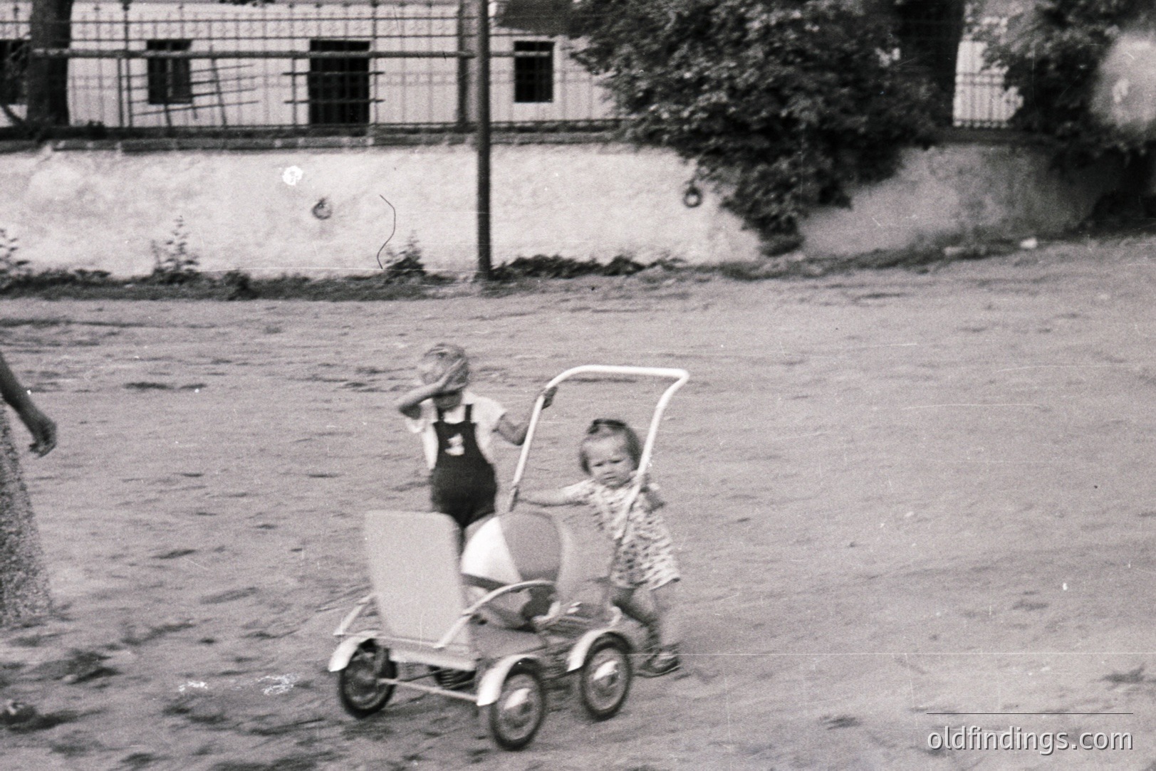 Two young children stand near a vintage metal tricycle; one playfully covers their face. The scene, likely a courtyard or yard, features a weathered wall and foliage. Architectural details suggest a European setting, potentially 1950s or 60s. Offers nostalgic appeal for design & historical research.