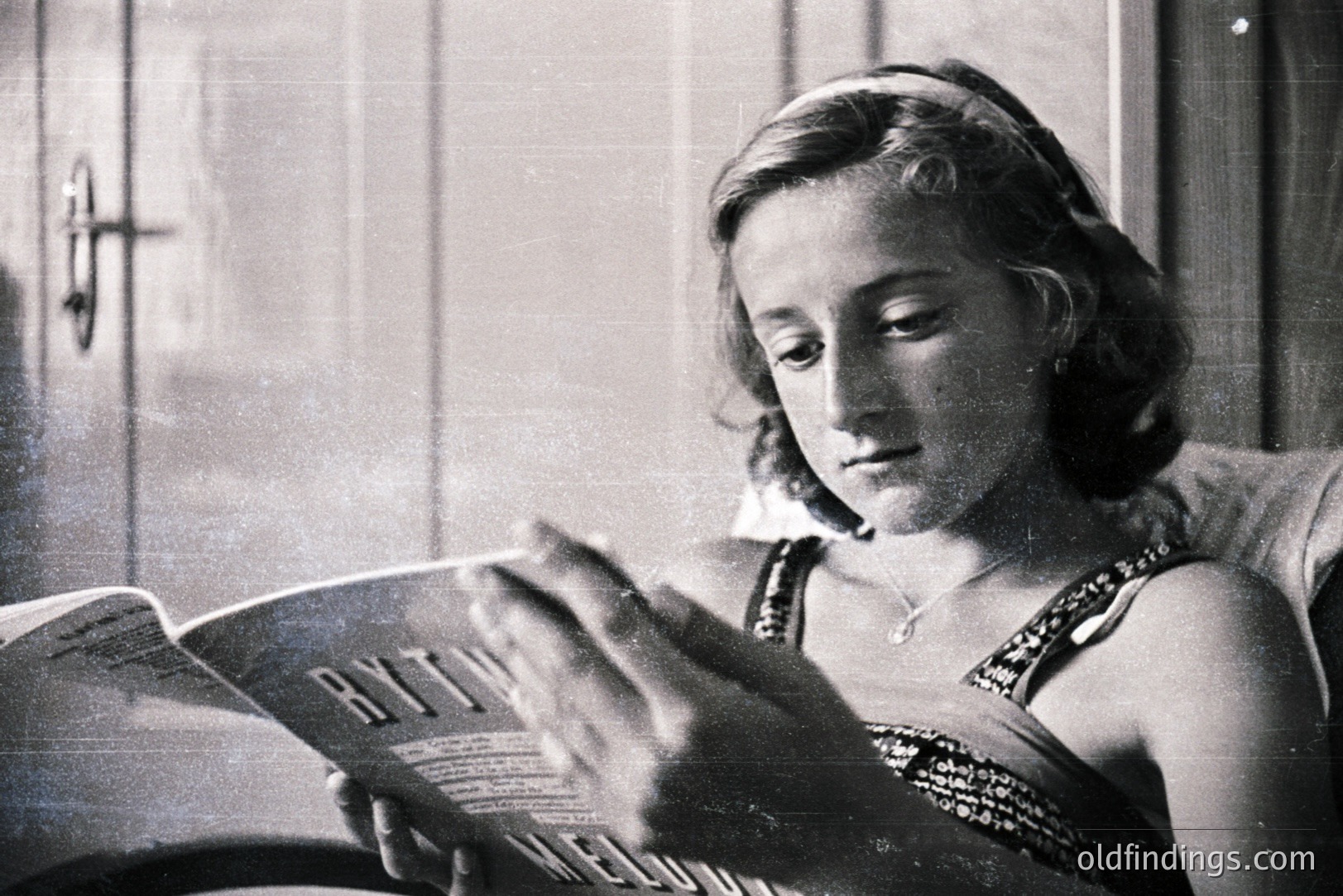 A young woman engrossed in a book, reclined on what appears to be an upholstered chair. She wears a decorative headband and a strapless dress, accessorized with a delicate necklace. Strong side lighting emphasizes her profile and the texture of the wooden wall in the background. Likely a portrait from the 1940s. Potentially useful for vintage design or advertising concepts.