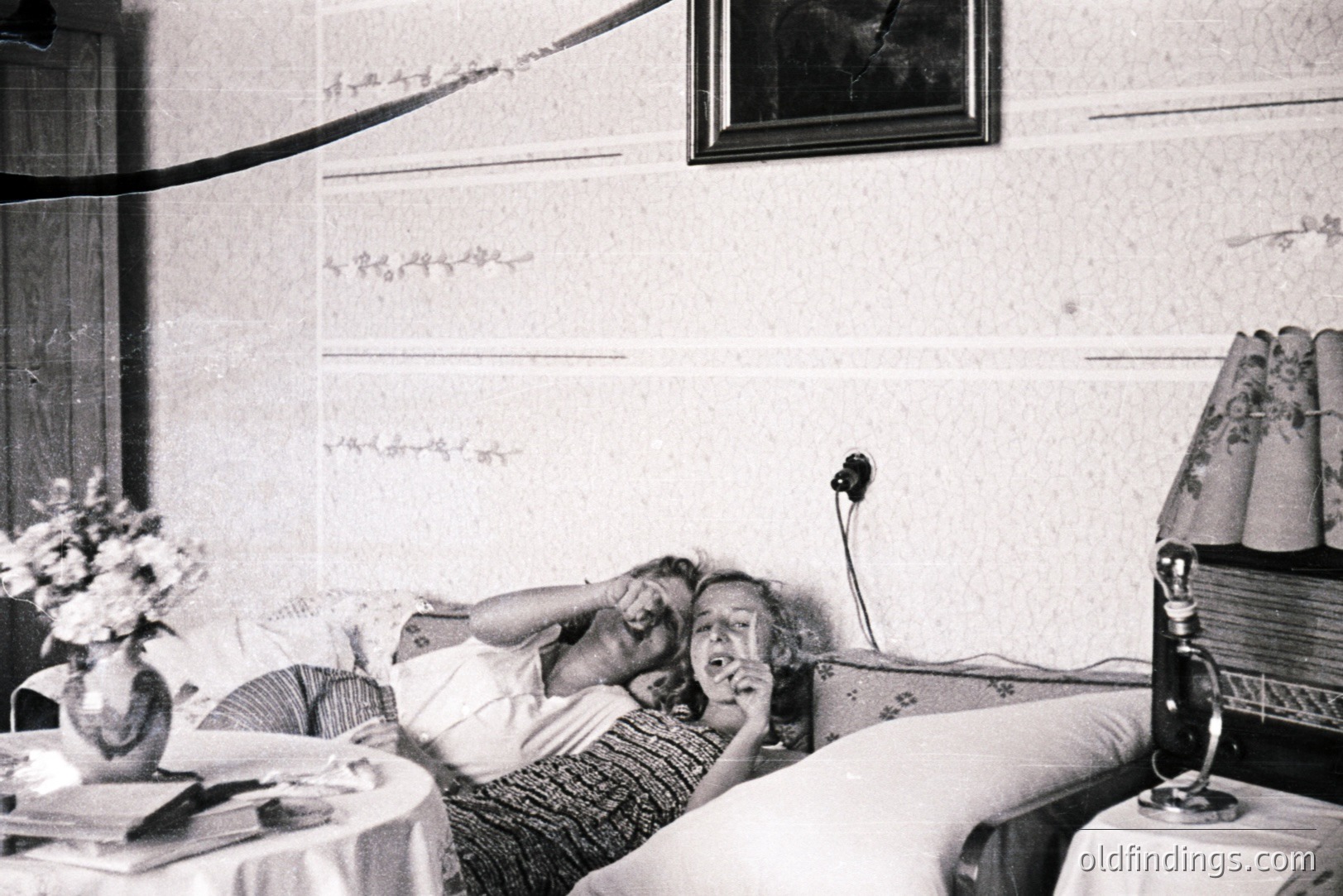 A young woman reclines on a bed, playfully gesturing with one hand near her face. A floral arrangement sits on a nearby table, along with books. The room’s wallpaper and patterned bedspread suggest a domestic interior, circa 1930s. A framed portrait hangs on the wall.