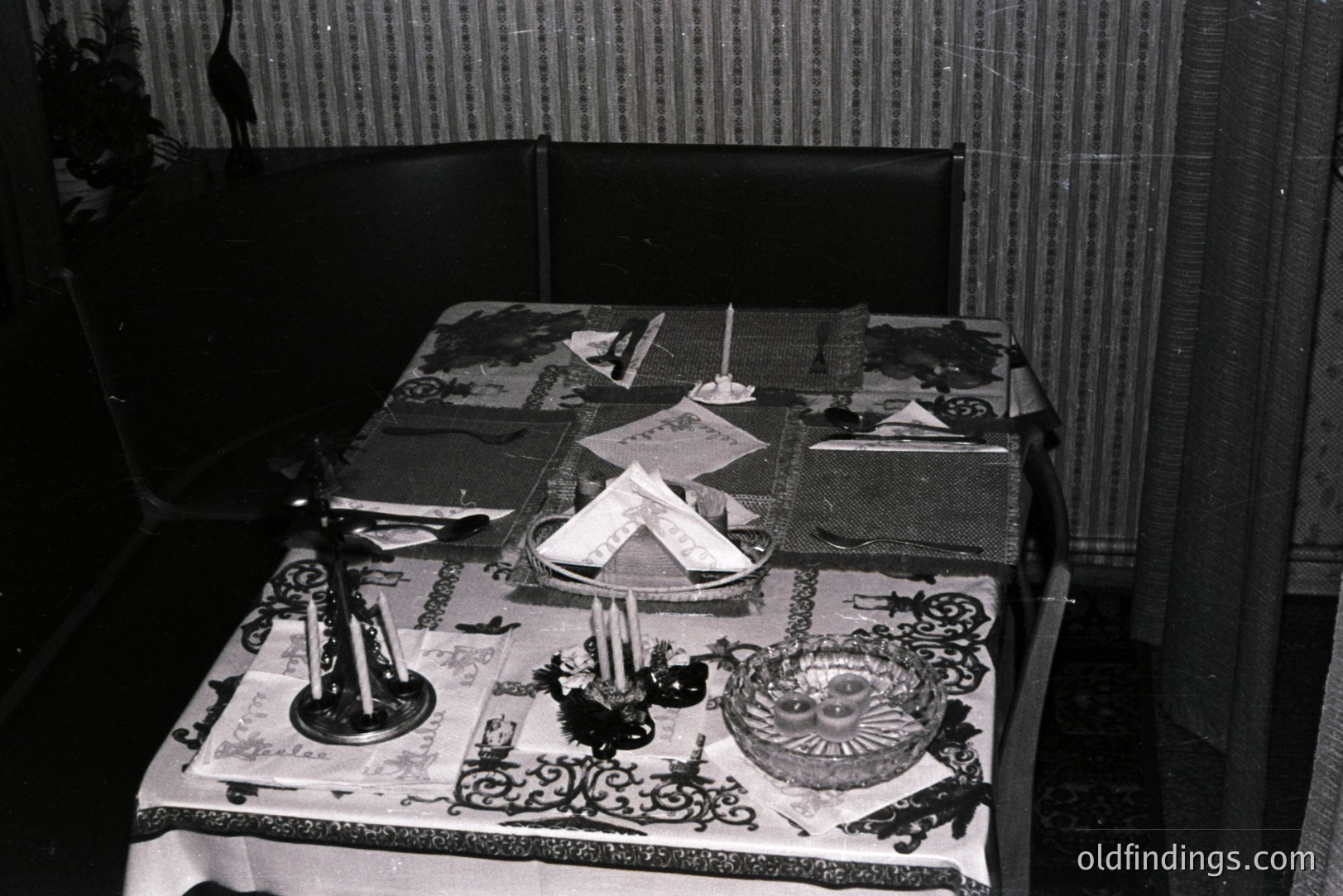 A table set with a patterned tablecloth, napkins, candle holders, and a decorative bowl. The backdrop includes a dark upholstered chair and textured wallpaper, typical of mid-century interior design. Likely 1960s-1970s home decor. A candid, domestic scene.