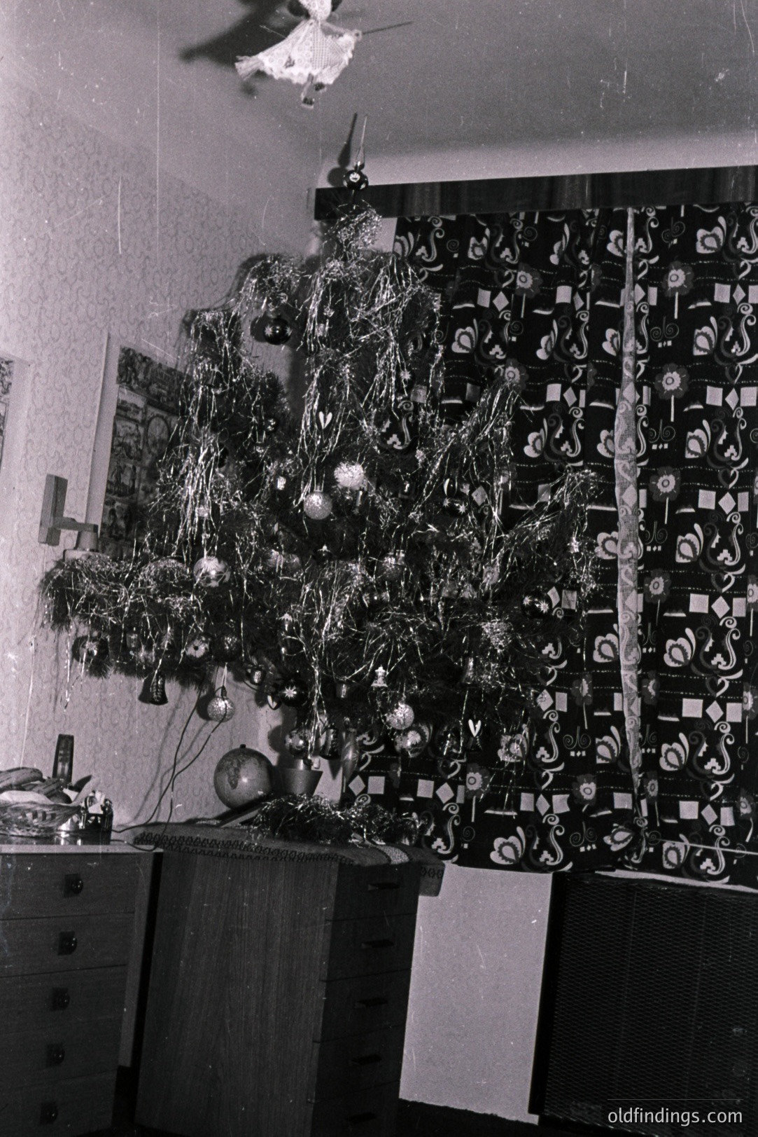 Interior view captures a sparsely decorated Christmas tree in a domestic setting. Ornaments, tinsel, and a ballerina figurine adorn the tree. A patterned curtain and dresser suggest a 1960s-70s aesthetic. Likely a family snapshot.