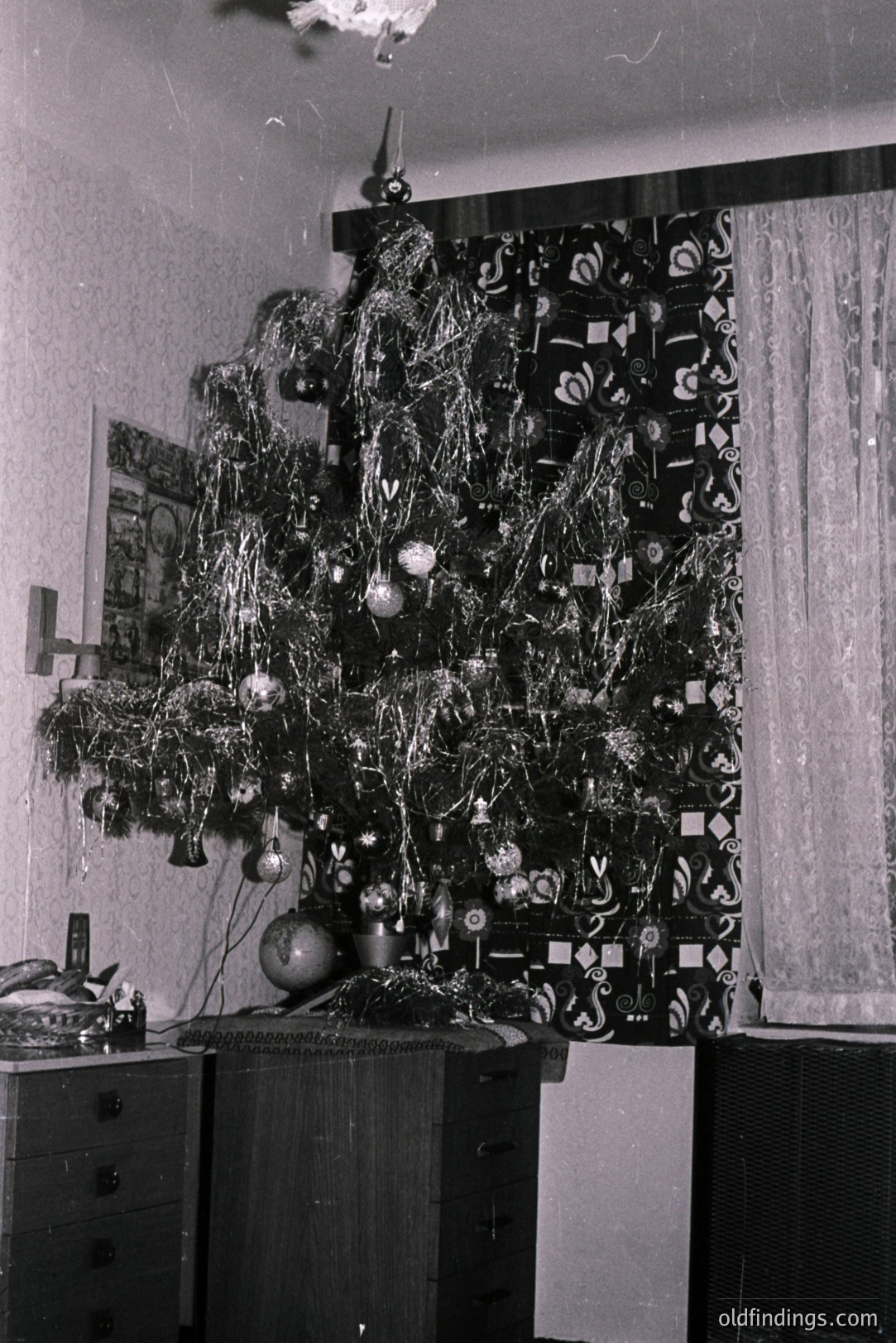 Interior scene showing a tinsel-covered artificial Christmas tree placed in front of a wall with patterned wallpaper and draped curtains. Dark wood furniture flanks the tree. Likely 1960s/70s decor. A vintage family snapshot hangs on the wall. Evokes nostalgic holiday charm.