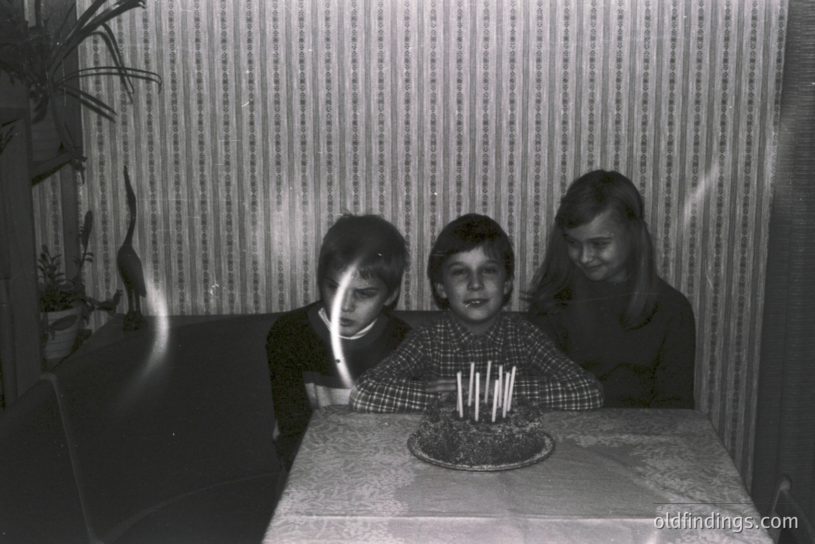 Three children gather around a birthday cake with numerous lit candles on a lace-covered table. Mid-1970s style wallpaper serves as the backdrop. Likely a family snapshot capturing a childhood celebration. The image exhibits a candid, home photography aesthetic.