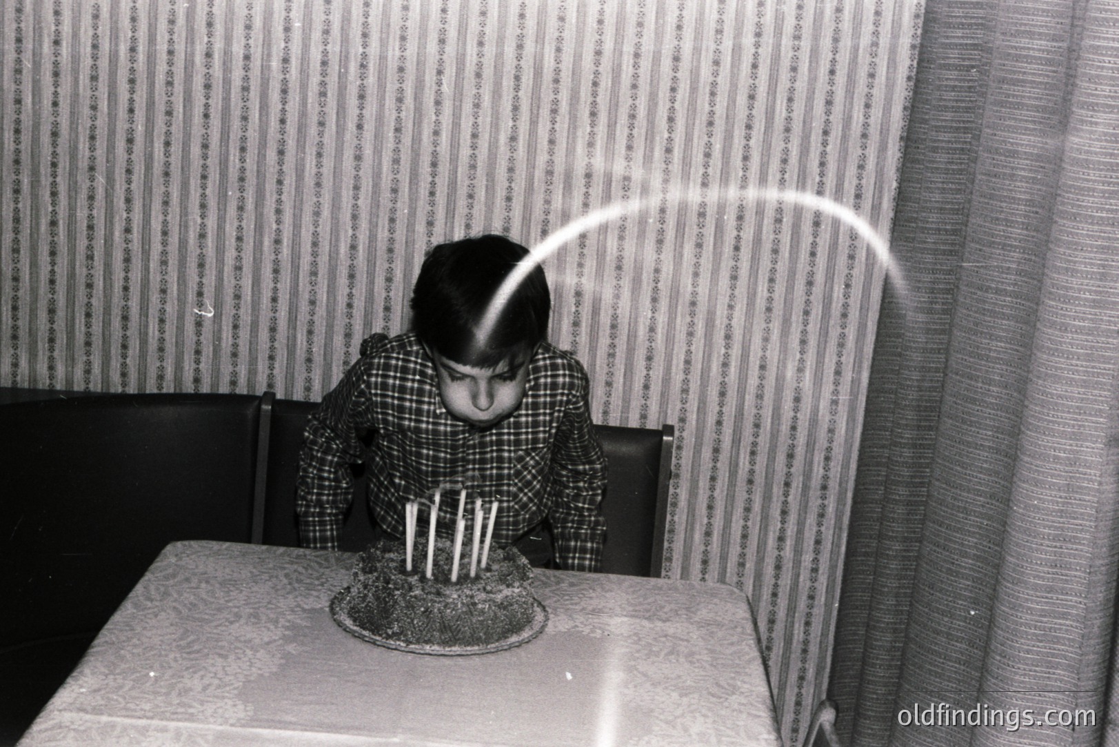 A young person blows out candles on a frosted cake, captured with a motion blur effect showing the exhalation. Seated at a covered table, backdrop reveals a striped wallpaper and heavy drapes. Likely a candid, informal portrait.