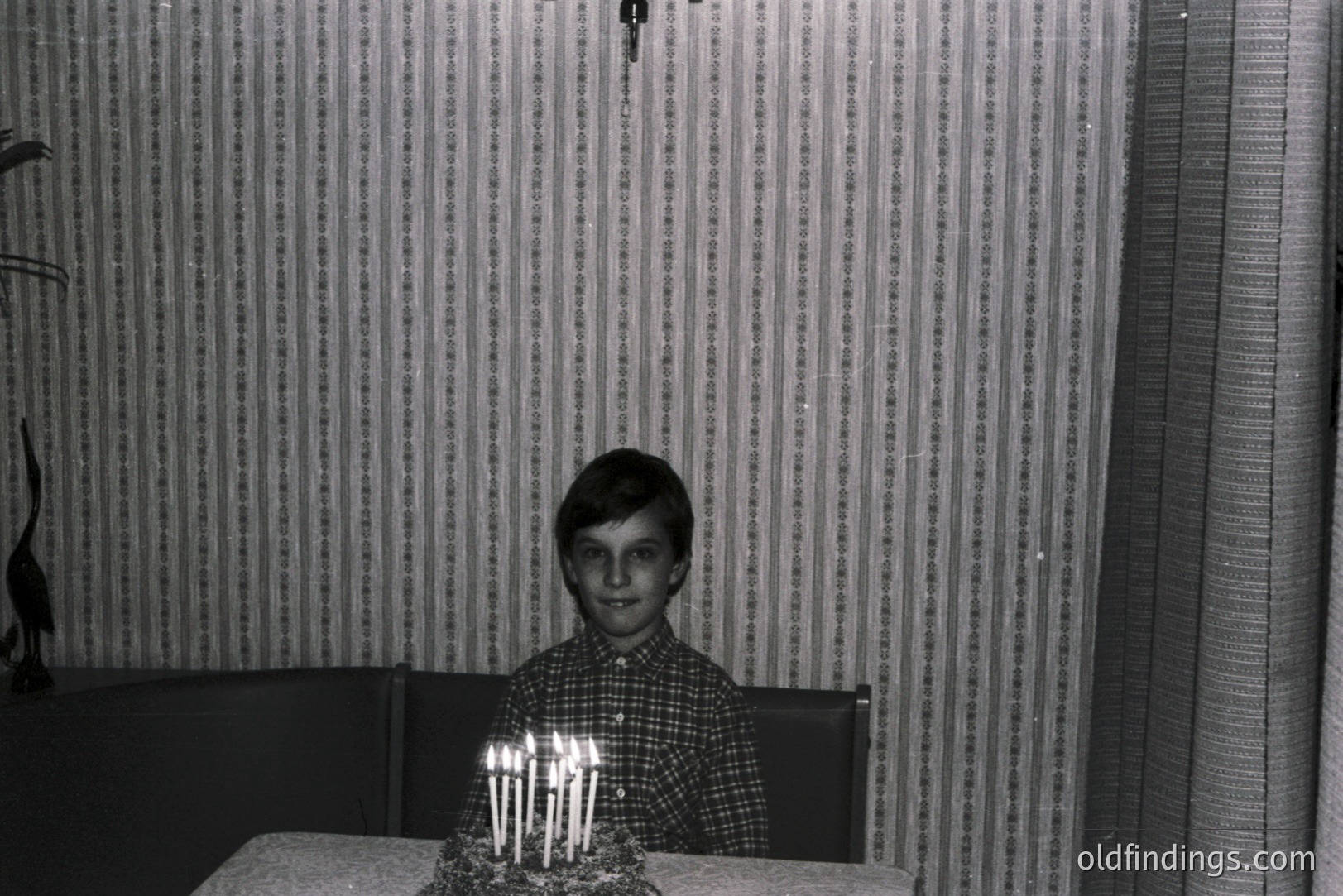A young boy sits at a table, a birthday cake adorned with numerous lit candles before him. The boy wears a patterned, button-down shirt. The background features textured wallpaper and a decorative sculpture. Likely a family snapshot, c. 1960s-1970s. Evokes nostalgic, domestic scenes.