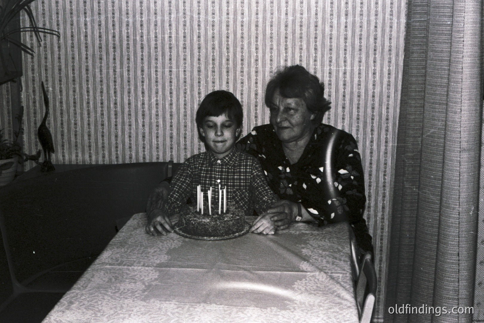 A young boy holds a birthday cake with four candles at a table covered in a lace tablecloth. A smiling elder woman stands beside him, both in a room featuring patterned wallpaper and a draped window. Likely a family snapshot, c. 1970s. Appears to be a candid moment.