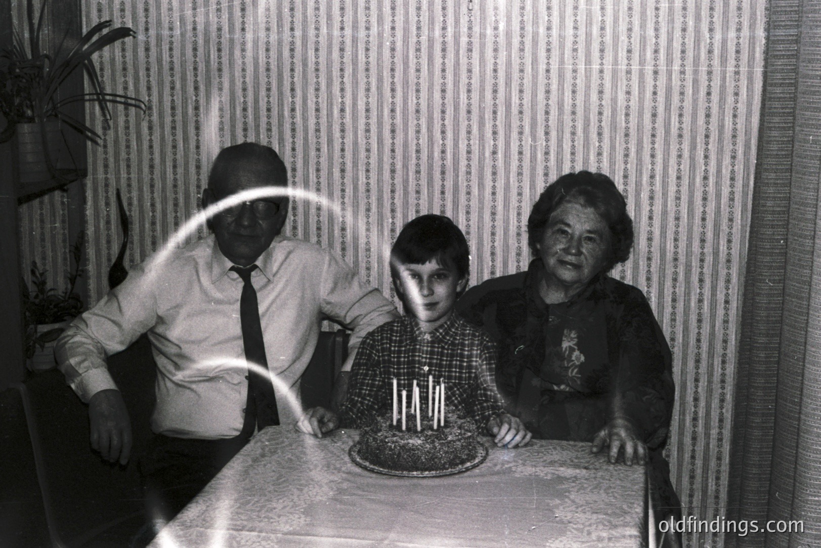A vintage black and white photo shows a boy celebrating a birthday with his grandparents seated on either side. A cake with lit candles sits on a tablecloth-covered table. Likely mid-1960s or 70s, capturing a domestic moment. The room's textured wallpaper and clothing styles are defining features.