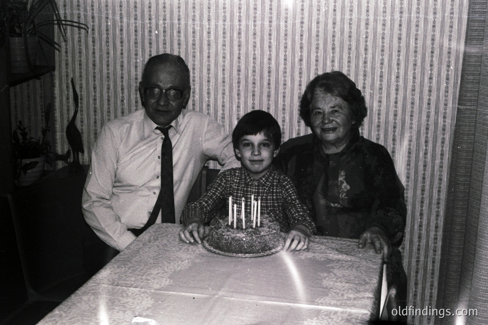 A boy stands proudly with a birthday cake, flanked by an elderly man and woman. The scene evokes a mid-century family celebration, likely 1960s-1970s. Note the patterned wallpaper and table cloth—typical domestic decor of the era. A cherished family moment captured.