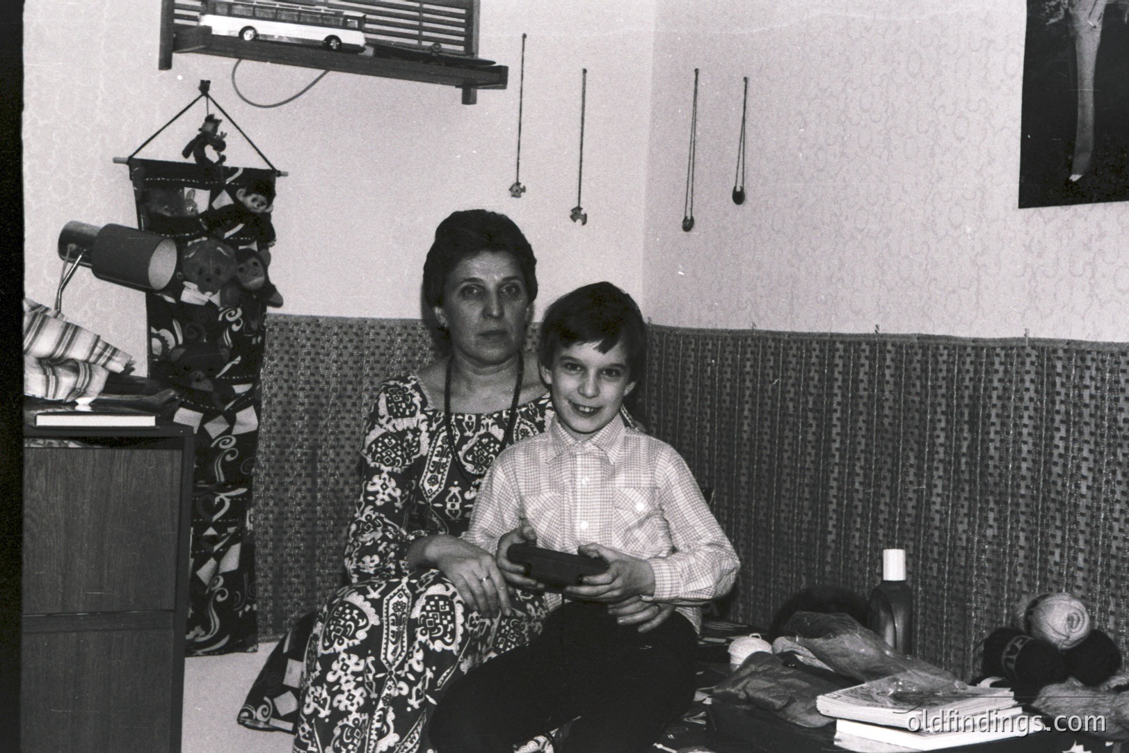 A mother and son pose in a richly decorated room, circa 1970s. The woman wears a patterned dress; the boy, a button-down shirt. Visible details: textured wallpaper, woven wall hanging, stacked books, & framed art. Evokes a sense of home life.