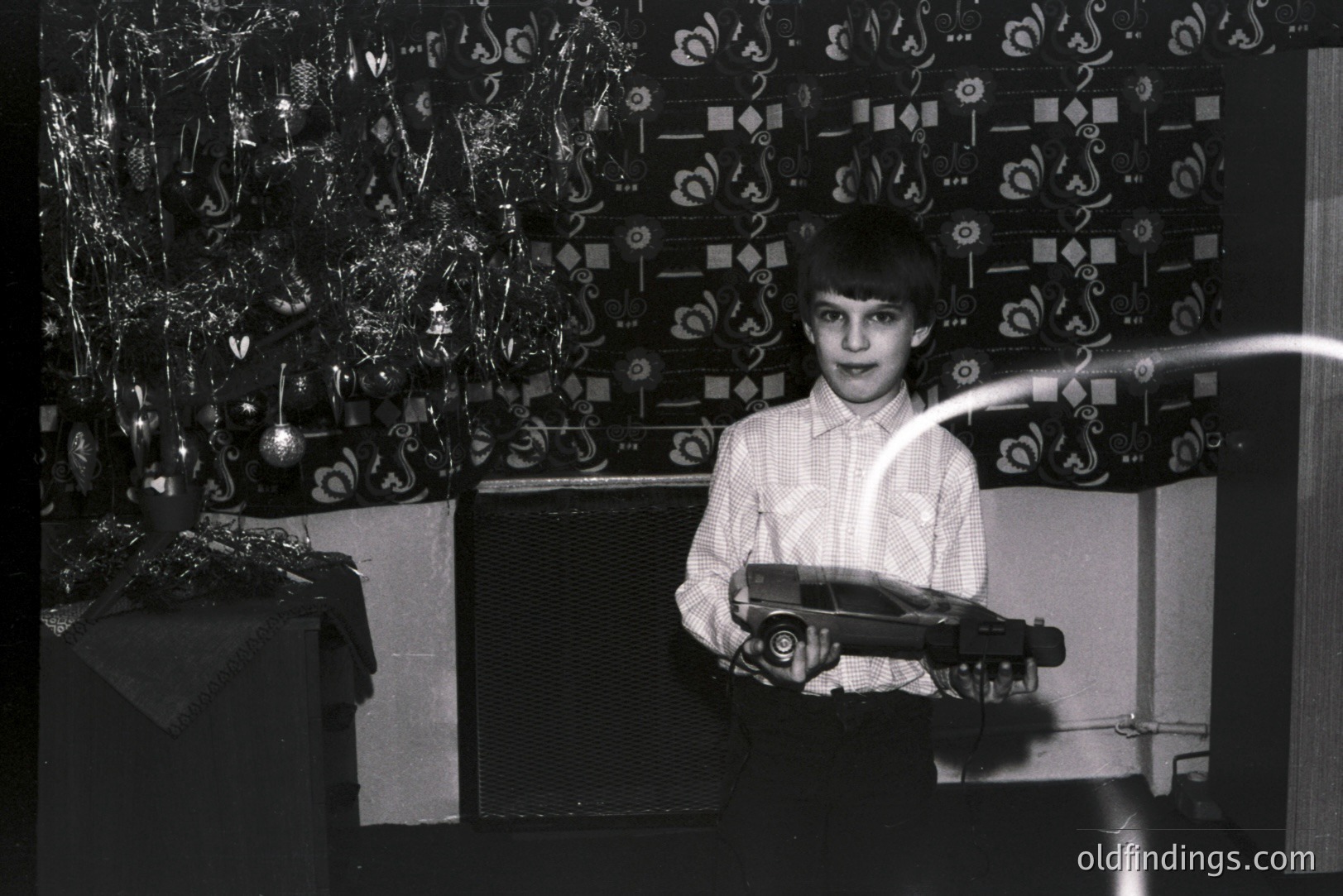 A young boy displays a toy car against a backdrop of a decorated Christmas tree and patterned curtains. The image, likely from the 1970s, utilizes long exposure to create light trails. The boy’s attire—a collared shirt and trousers—indicates a mid-century aesthetic.