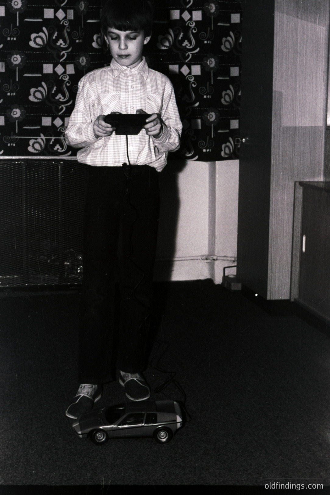 A young boy stands operating a toy radio-controlled car indoors. He wears a button-down shirt and wide-legged trousers, typical of the 1970s. The room features patterned wallpaper and a radiator, indicative of mid-century domestic design. A nostalgic glimpse of childhood play.