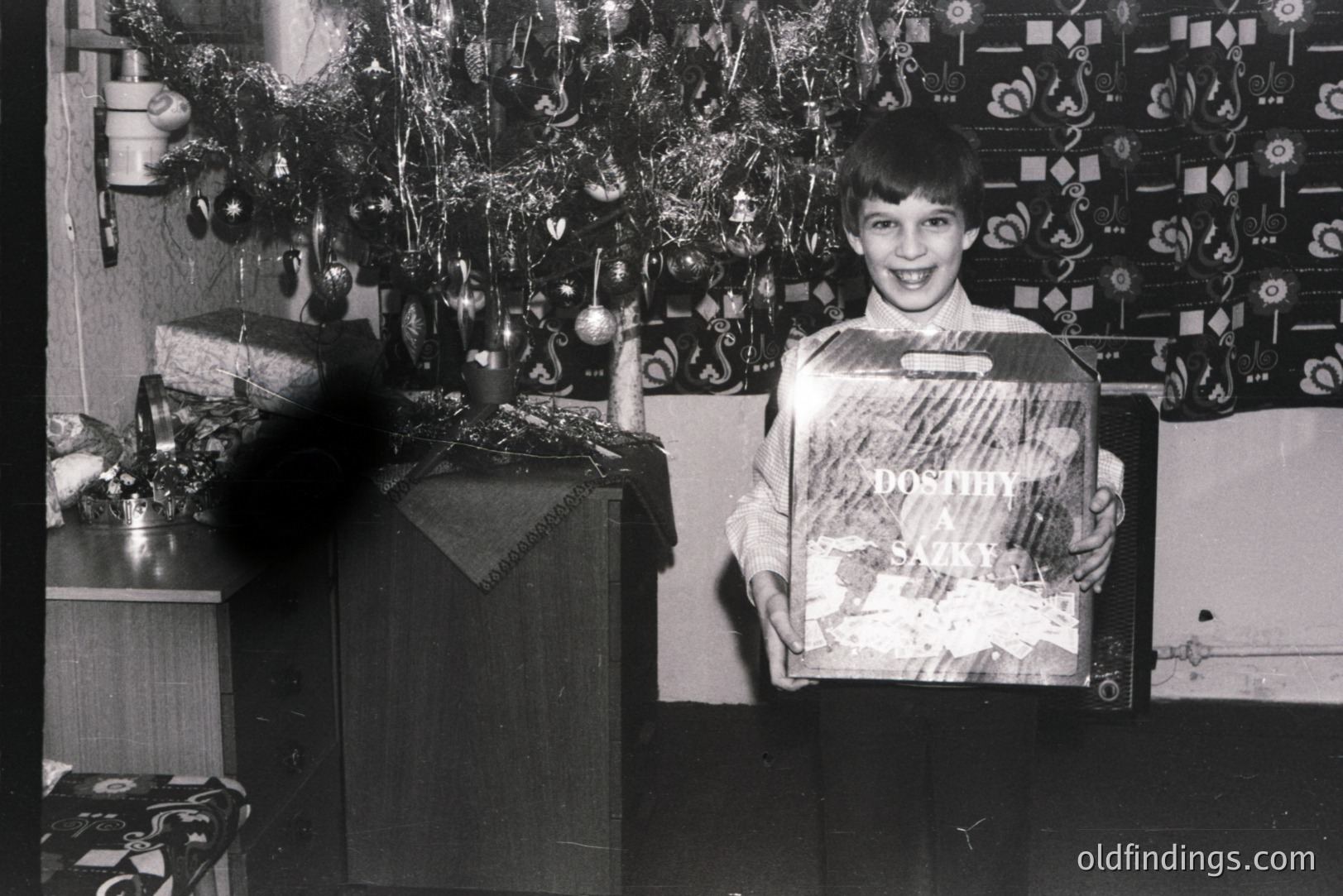 A young boy stands proudly, displaying a "Dostih a Sazky" branded gift box beneath a decorated Christmas tree. Mid-century modern furniture and patterned wallpaper suggest a 1960s-70s interior. Likely a family photograph capturing holiday joy and consumer culture.