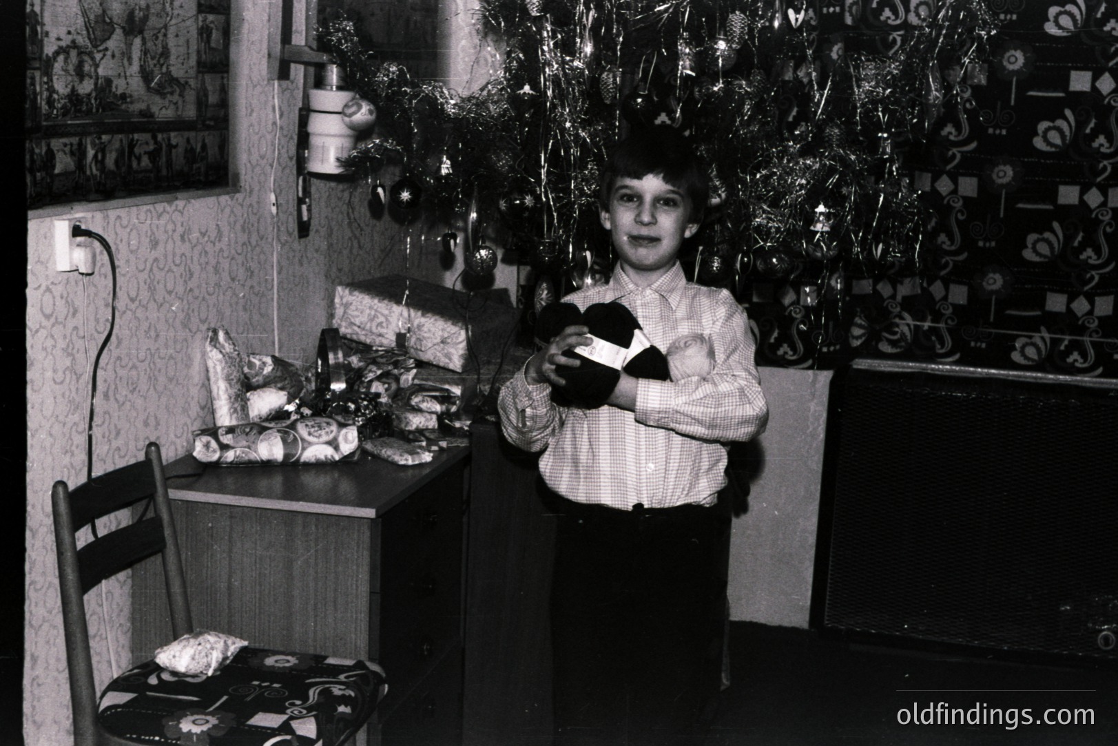 A young boy stands proudly holding a wrapped gift, posed in front of a decorated Christmas tree. Visible are gift packages, a patterned armchair, and textured wallpaper, indicative of a mid-century interior. Likely a family snapshot from the 1960s or 1970s.